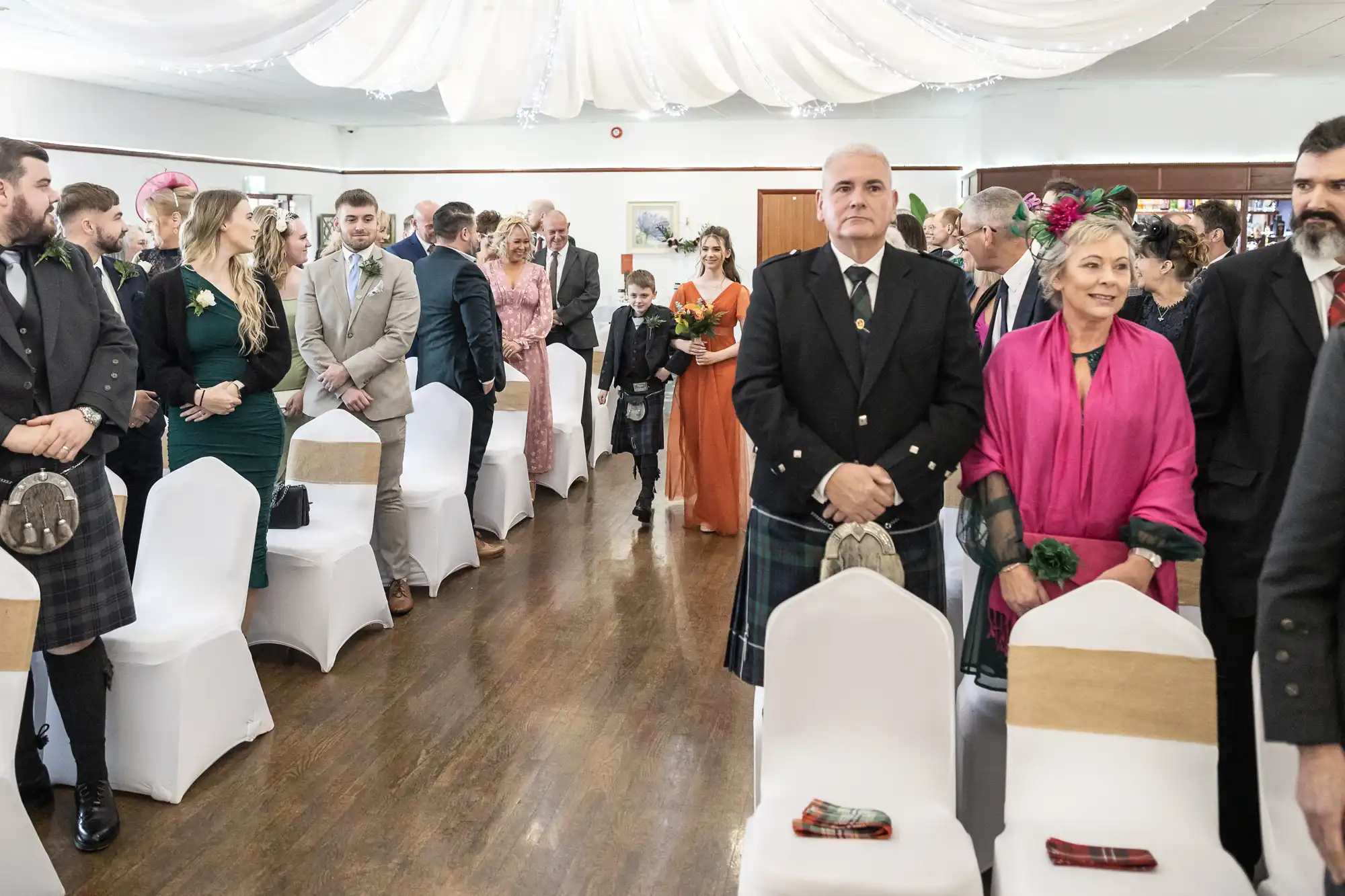 Wedding guests in formal attire stand in two rows beside white-covered chairs in a decorated hall. An older man in a dark kilt and an older woman in a bright pink shawl stand at the front. In the aisle, a woman in orange leads a young boy forwards.