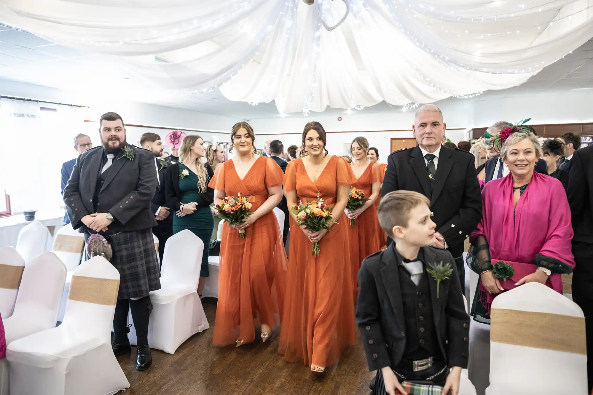 Three bridesmaids in matching orange dresses walk down the aisle holding bouquets. Guests sit on either side; some are standing and smiling, dressed formally. The room has draped white fabric and fairy lights hanging from the ceiling.