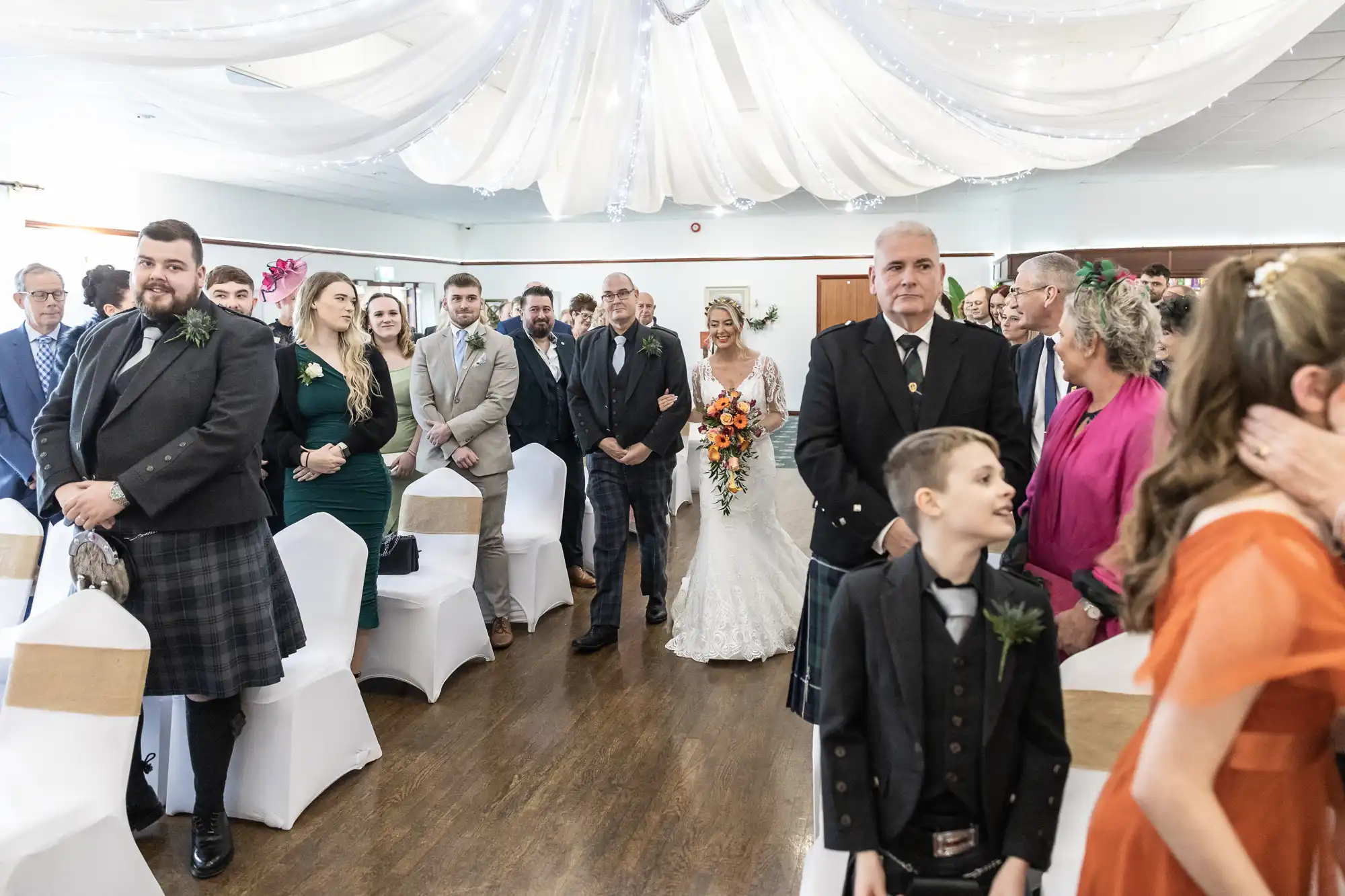 A bride in a white lace dress walks down the aisle with an older man in a kilt. Guests in formal attire, some in kilts, stand on either side smiling. White drapes and fairy lights hang from the ceiling in the decorated wedding venue.