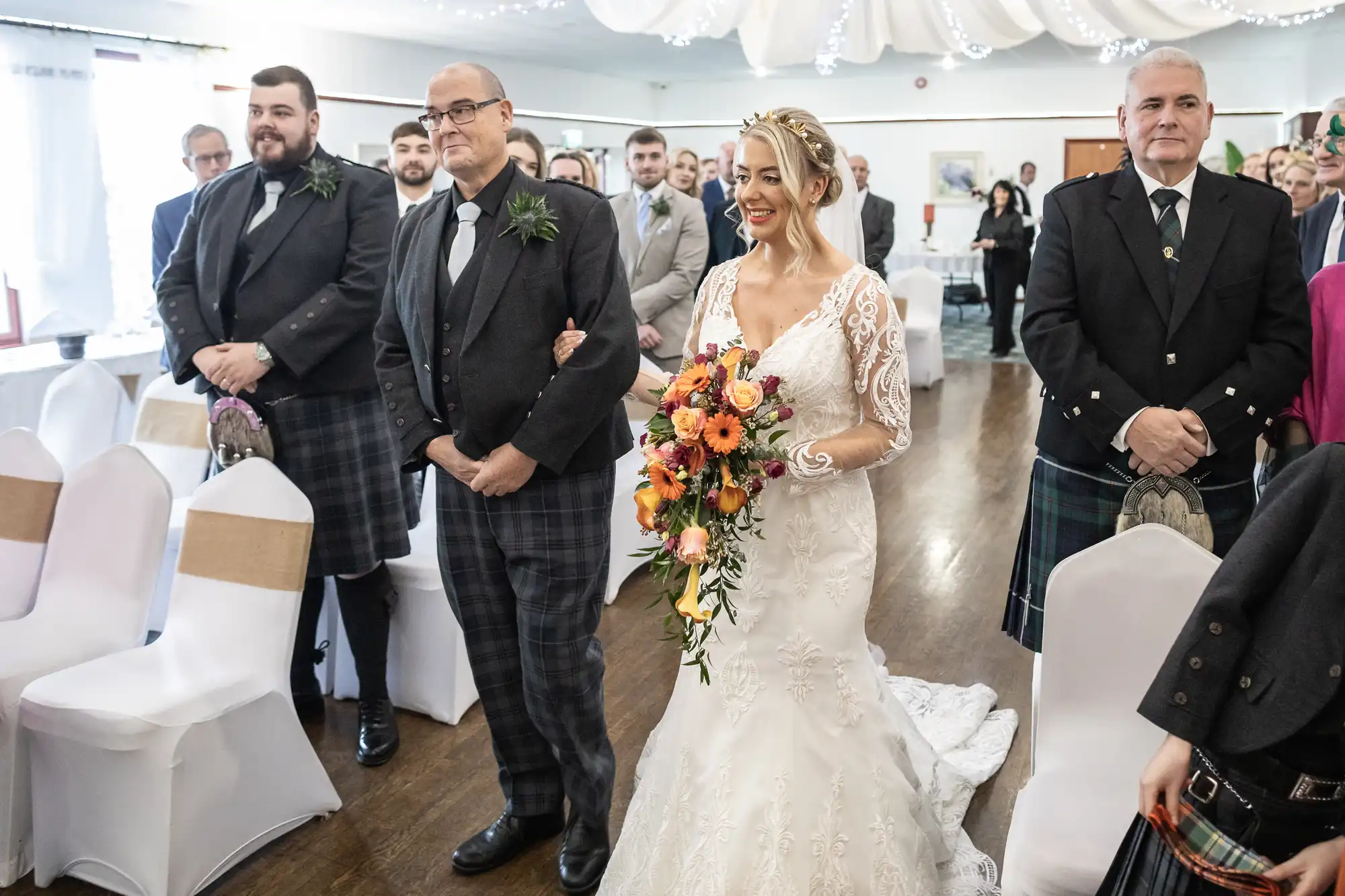 A bride in a white lace dress and veil holds a colourful bouquet while walking down the aisle with an older man in a suit and kilt. Guests, some in kilts, watch them in a decorated indoor venue with white chairs and flower arrangements.