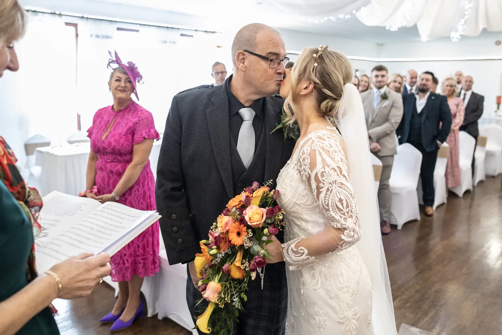 A bride in a long-sleeved lace gown and veil kisses an older man in a suit, likely her father, whilst holding a vibrant bouquet. Guests in formal attire watch and smile from their seats in a bright wedding ceremony room.