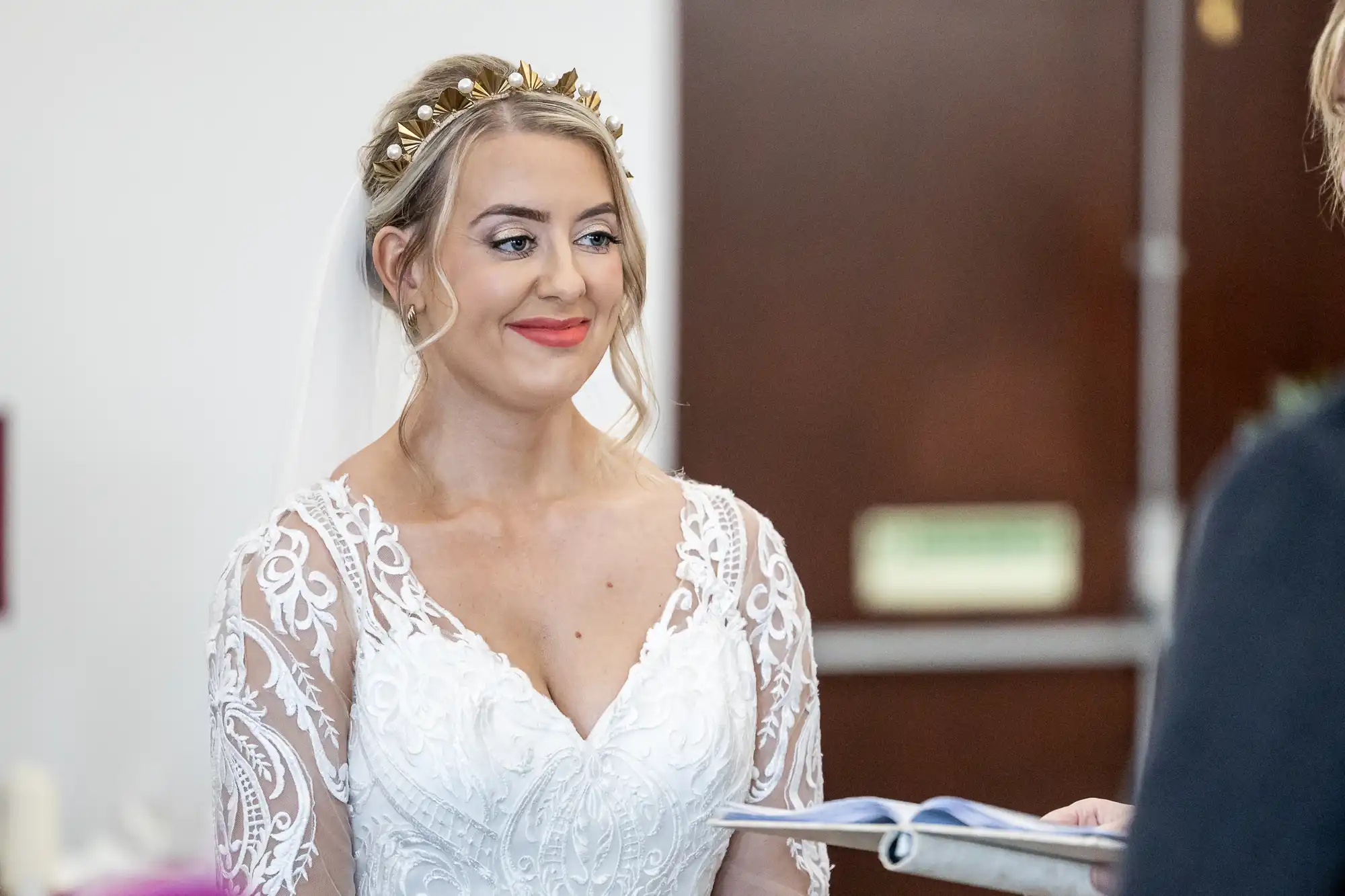 A bride in a white lace wedding dress and gold leaf headpiece smiles whilst looking at someone off-camera. She stands indoors, holding a booklet, with blurred doors and a sign in the background.