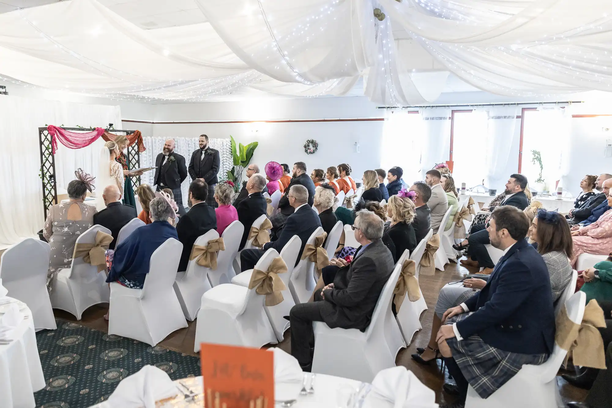A wedding ceremony indoors with guests seated on white chairs with gold bows, facing a couple and celebrant at the altar. The room is decorated with white draped fabric and fairy lights on the ceiling, and people wear formal and colourful attire.