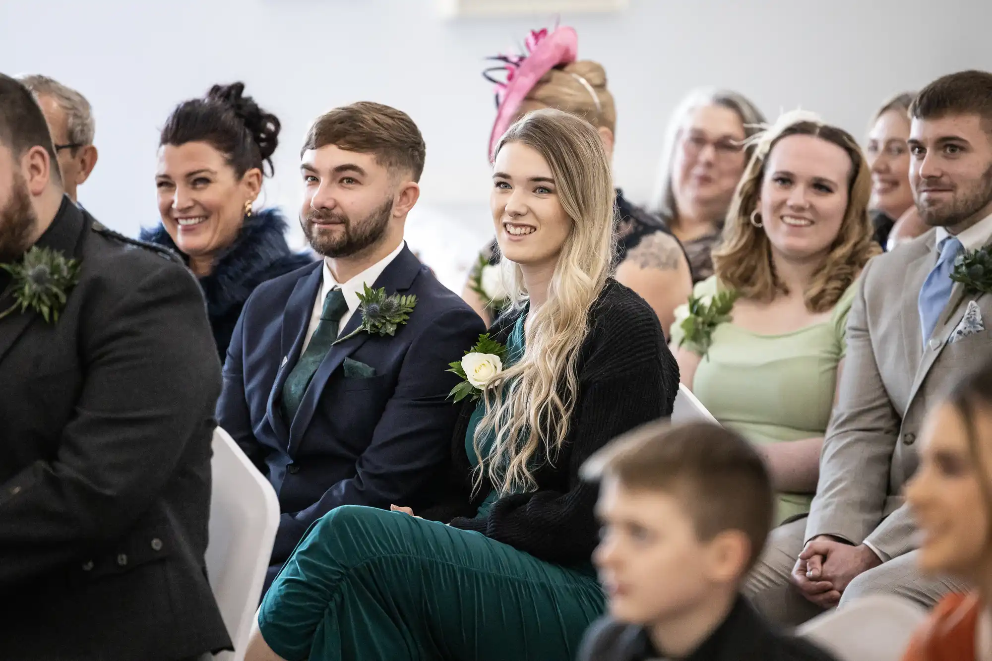 A group of well-dressed wedding guests sit in rows, smiling and attentively watching something off-camera. The focus is on a young man in a navy suit and a young woman in a green dress with long blonde hair holding a white rose. Other guests are behind them.