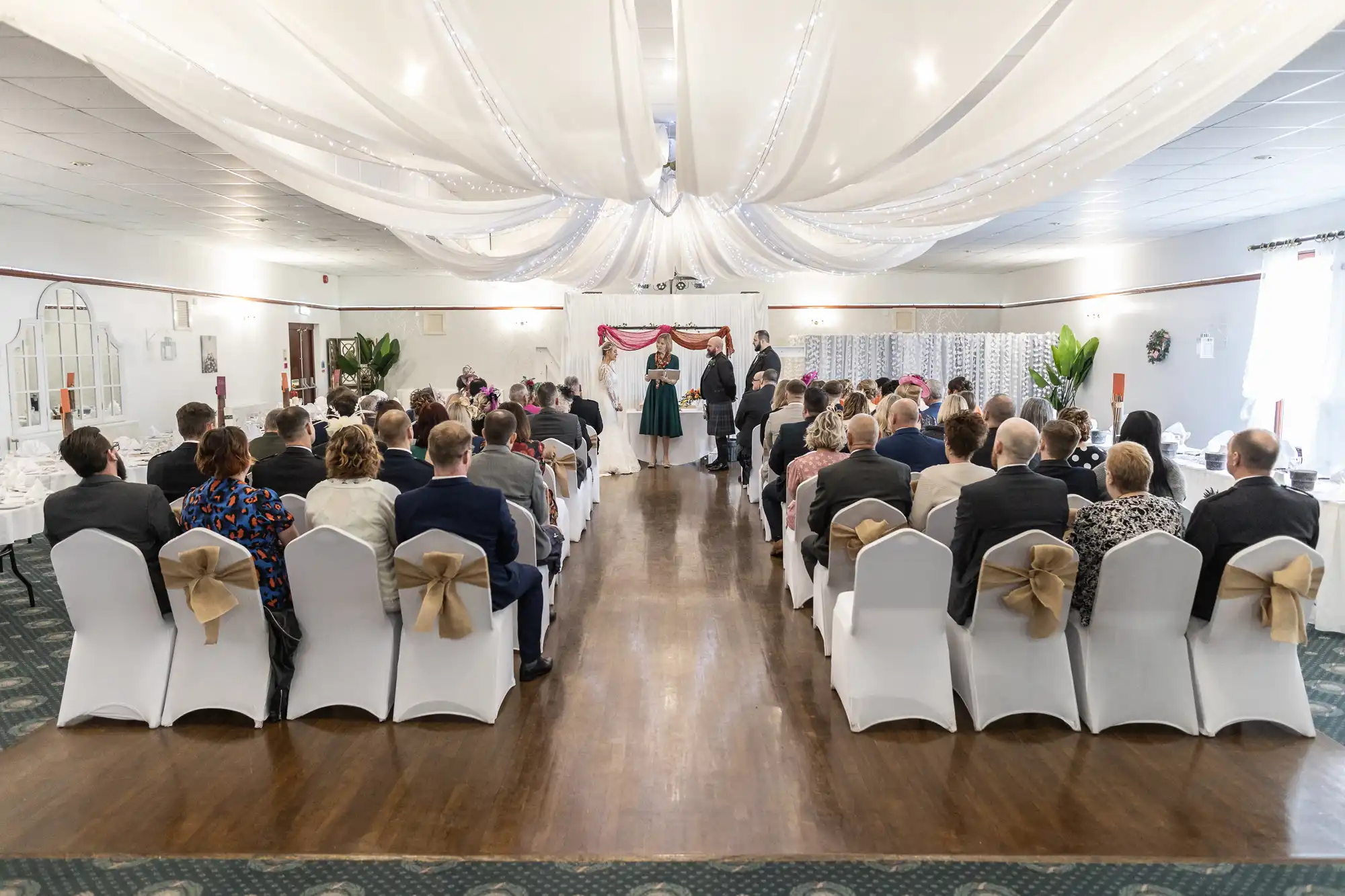 A wedding ceremony in a decorated hall with draped white fabric overhead. Guests sit on chairs covered in white cloth with tan bows, facing an aisle leading to a couple and celebrant at a small altar, with people seated on both sides watching.