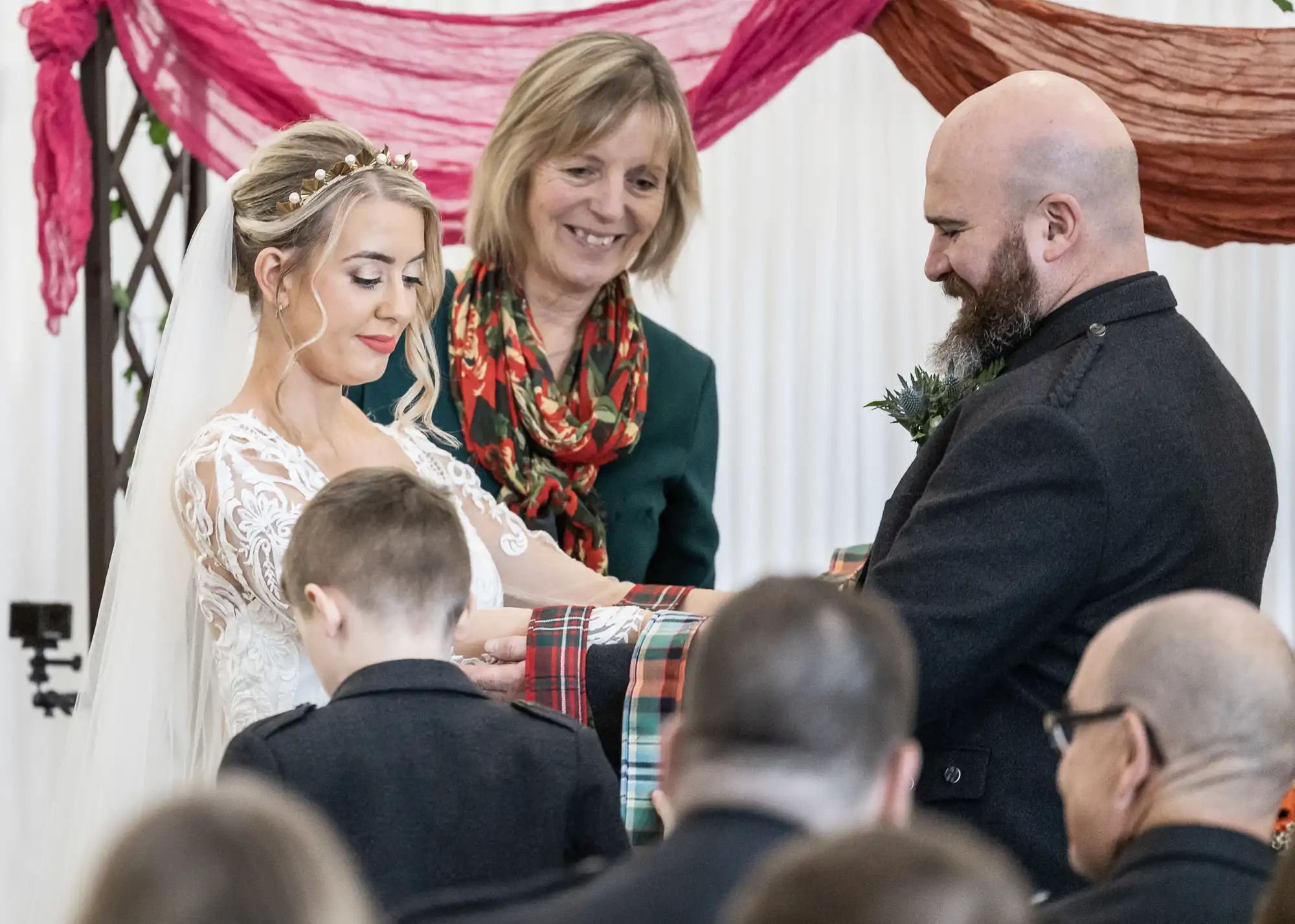 A bride and groom stand facing each other, smiling, as their hands are wrapped in tartan cloth by a woman during a wedding ceremony. The bride wears a white lace dress and the groom wears dark attire. Guests look on; pink and red fabric drapes in the background.
