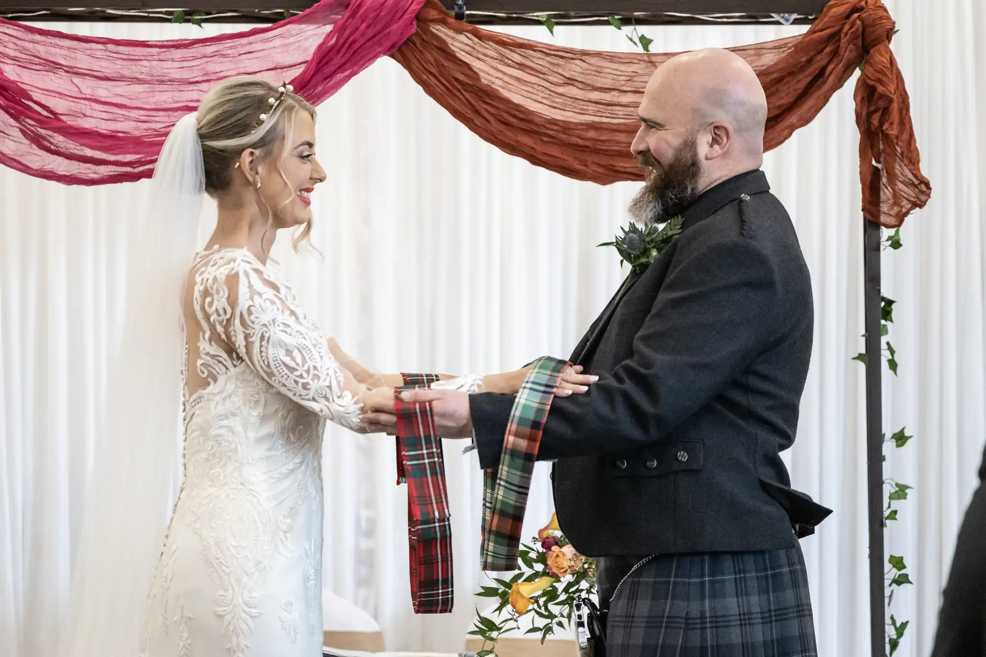 A bride in a white lace gown and a groom in a Scottish kilt smile at each other during their wedding ceremony, holding tartan ribbons together in a handfasting ritual. Colourful fabric drapes hang behind them.