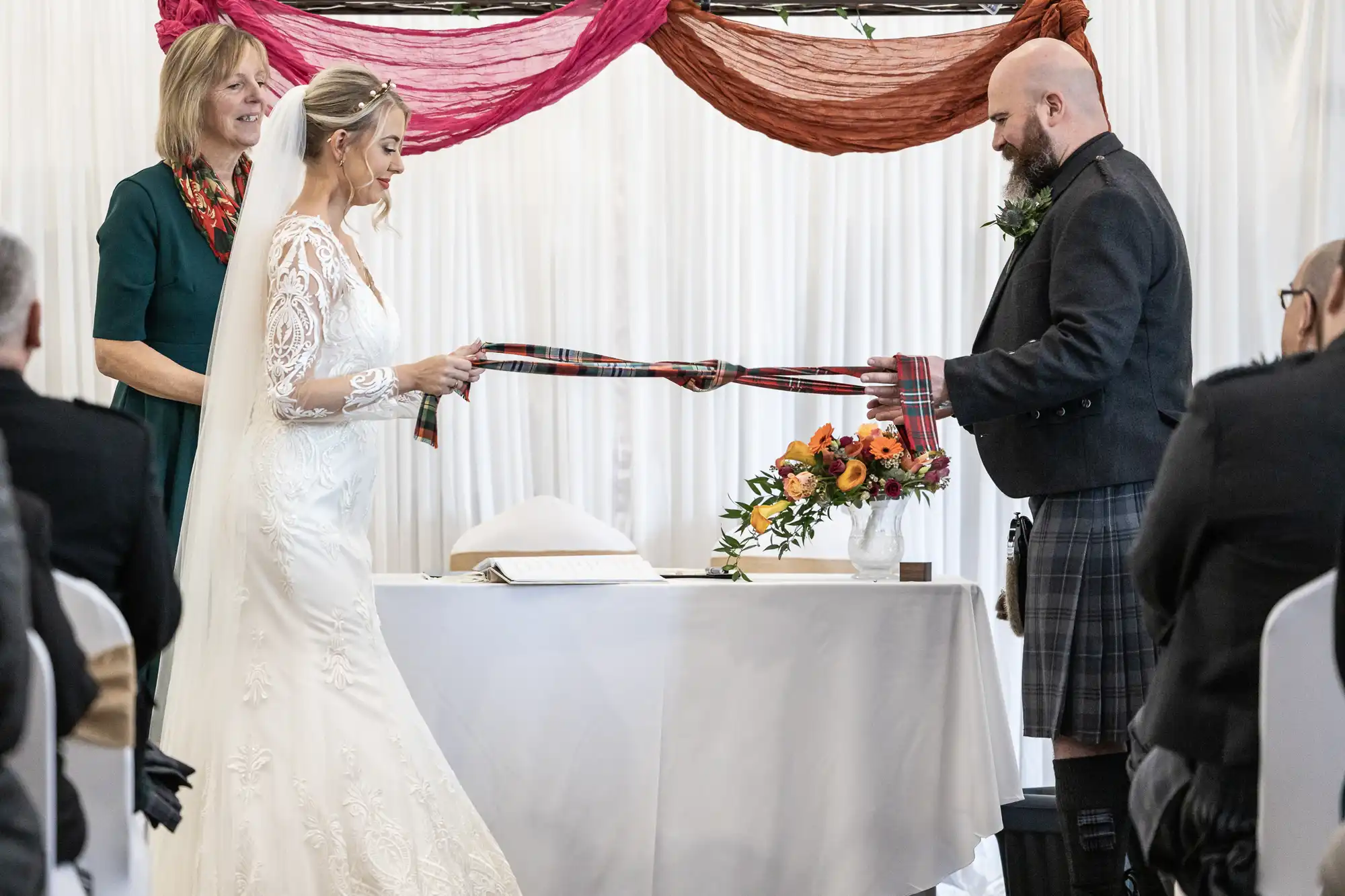 A bride in a white lace gown and a groom in a kilt perform a handfasting ceremony, holding tartan ribbons together as an officiant smiles beside them. They stand by a table with a floral arrangement in a bright, decorated setting.