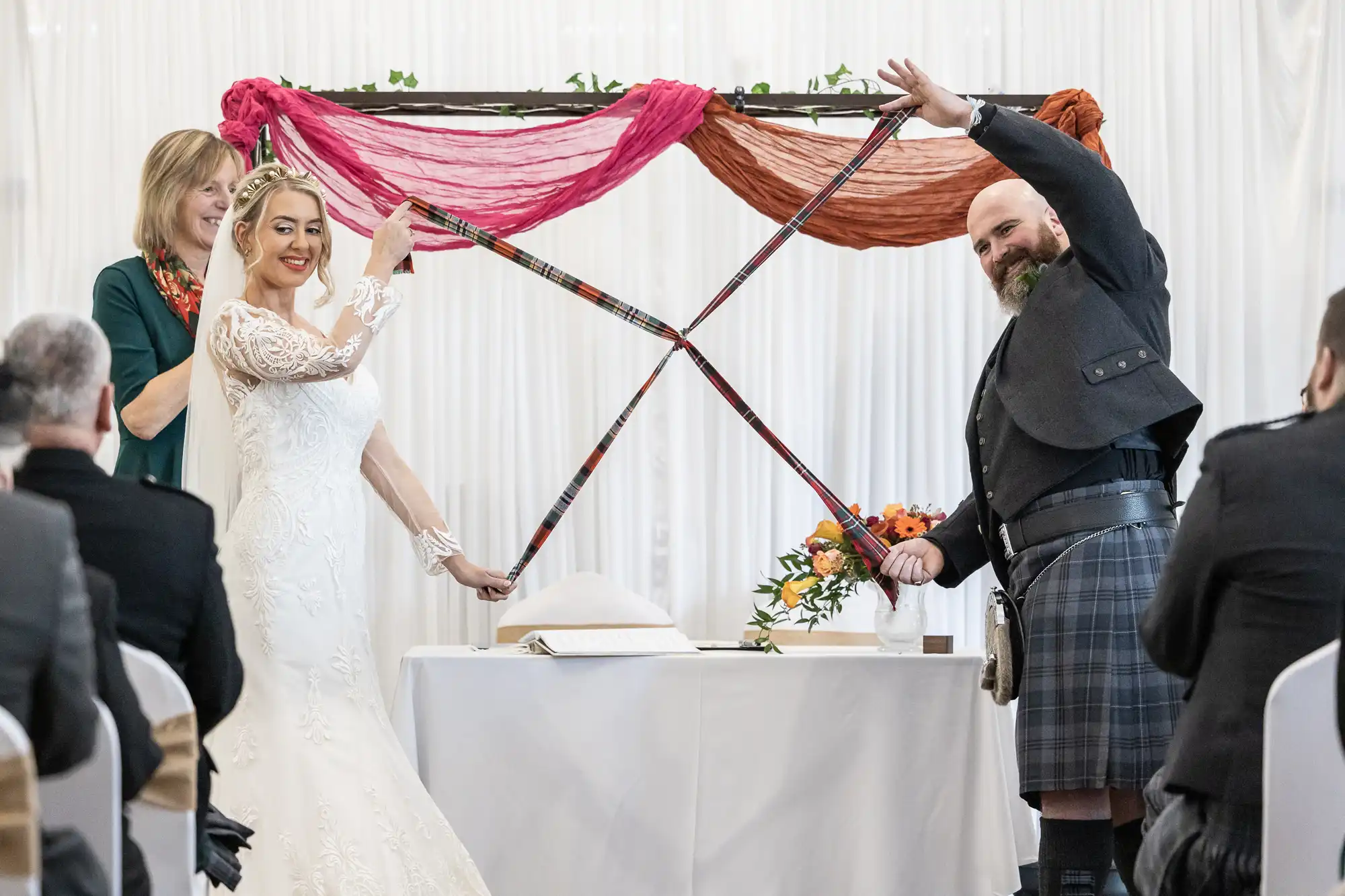 A bride in a white lace dress and a groom in a kilt hold colourful tartan ribbons, crossing them joyfully during a wedding ceremony. An officiant stands behind them, smiling. Guests watch as vibrant fabric decorates the background.