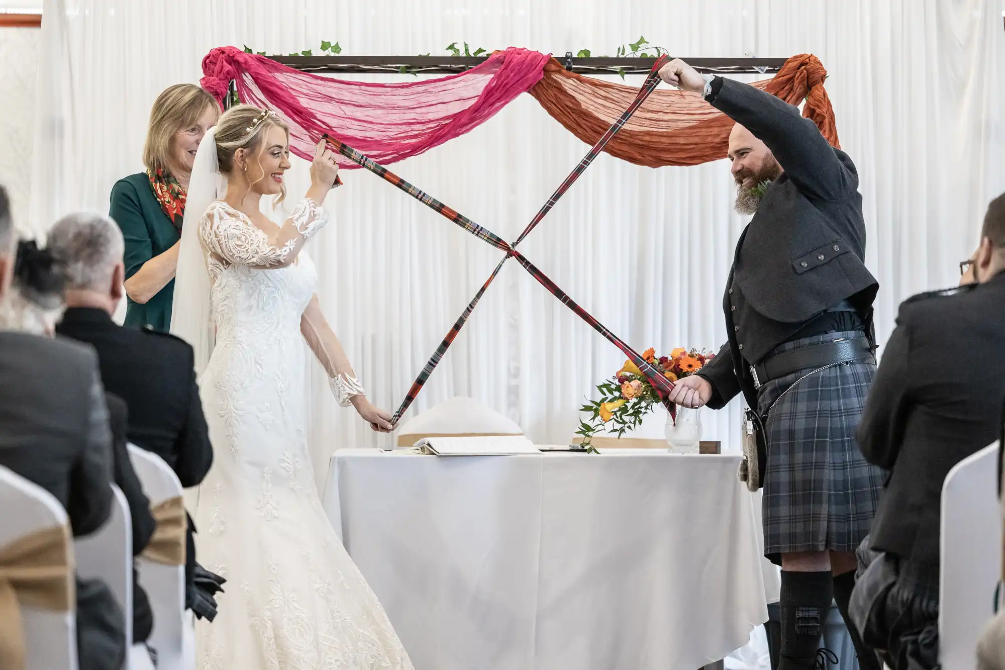 A bride in a lace dress and a groom in a kilt touch swords above an altar during a wedding ceremony, overseen by an officiant. Draped fabric decorates the background, with guests seated in the foreground watching the ritual.