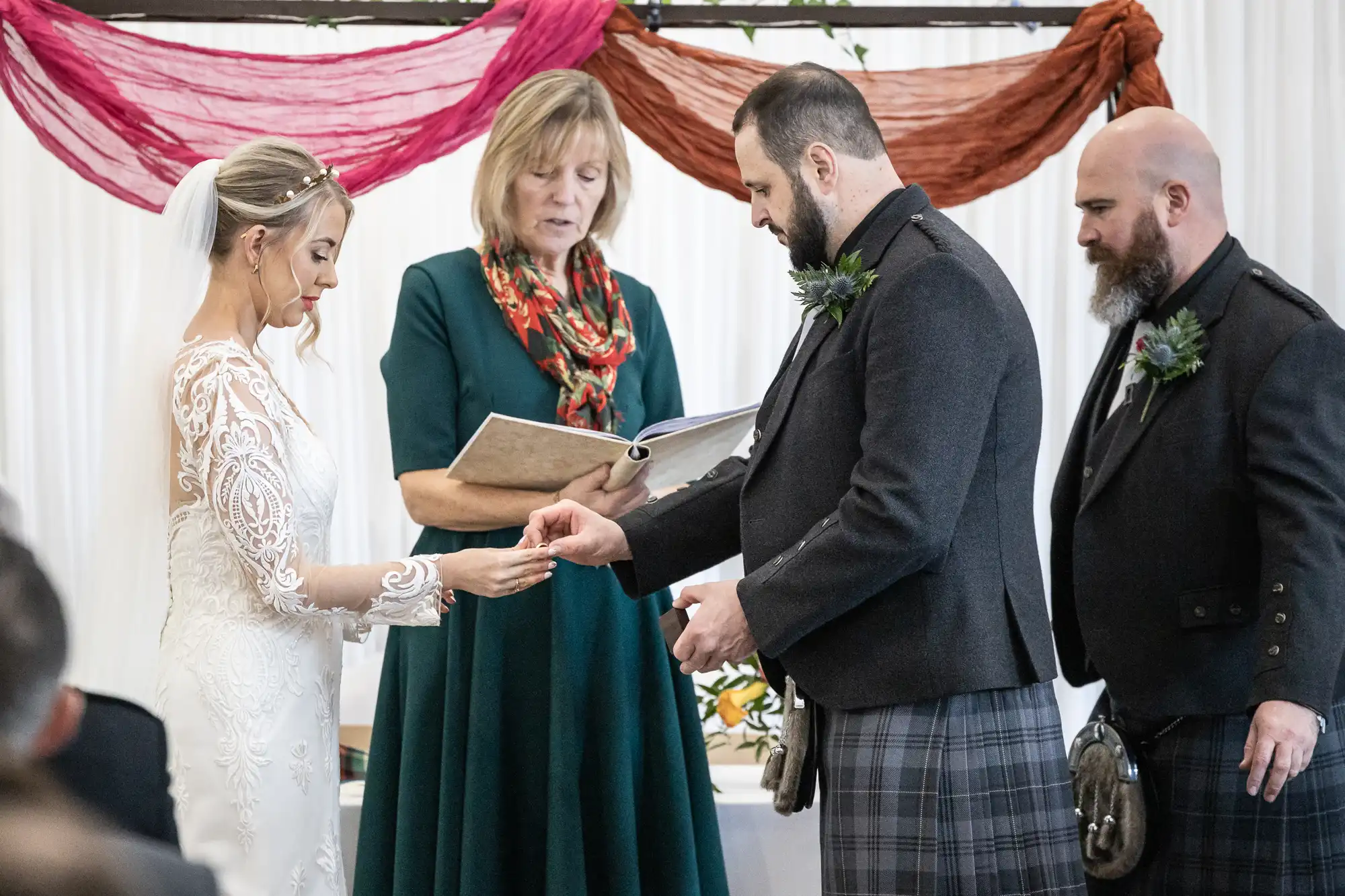 A bride and groom exchange rings during a wedding ceremony. The officiant stands between them, reading from a folder, while another man in a kilt stands nearby. Decorative drapes hang in the background. Everyone is dressed formally.