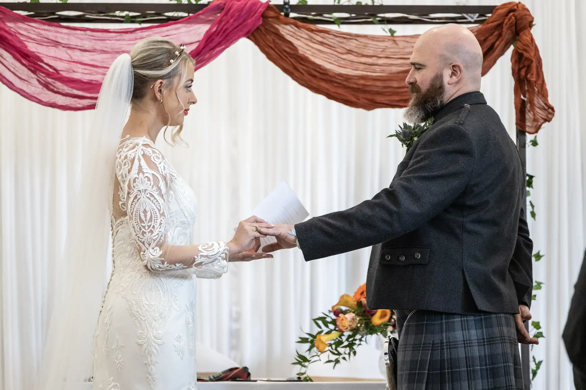 A bride in a lace wedding dress and veil holds hands with a groom in a dark jacket and checked kilt. They face each other, exchanging vows, with colourful drapery and a floral arrangement in the background.