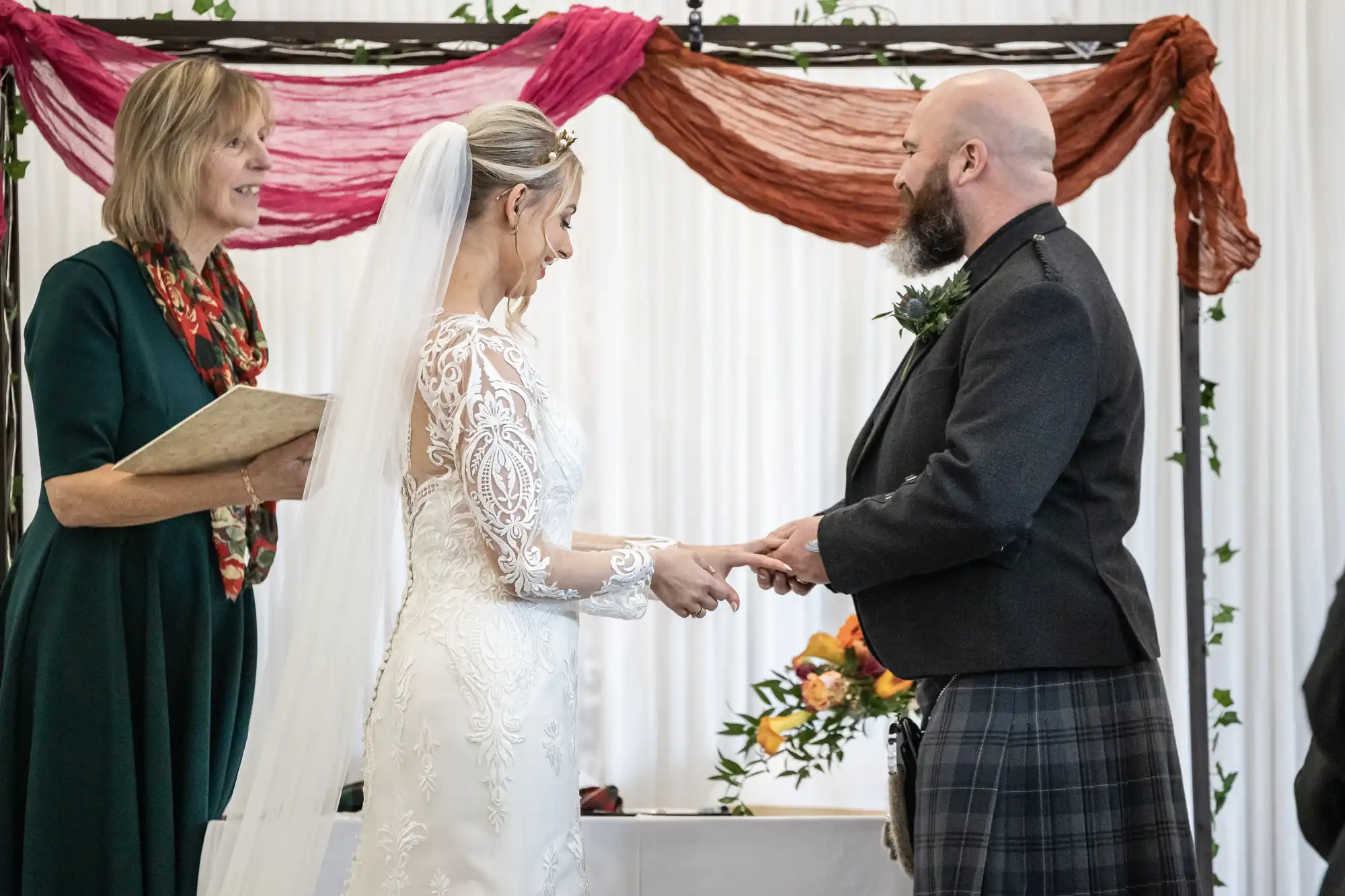 A bride in a white lace gown and veil exchanges rings with a smiling groom in a dark jacket and kilt. An officiant in a green dress stands beside them holding a book. Behind them are draped red and orange fabrics and floral decorations.