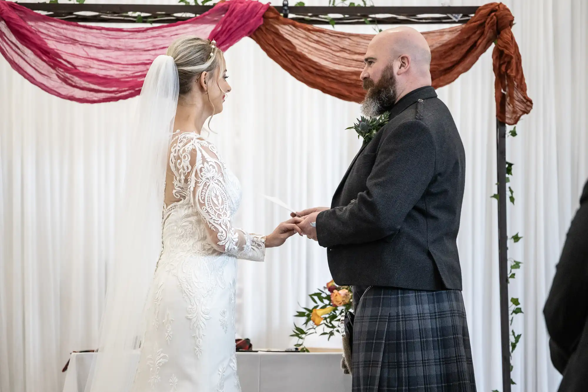 A bride in a long-sleeved lace dress and veil faces a groom in a dark jacket and tartan kilt. They hold hands, exchanging vows under an arch draped with red and brown fabric, with flowers on a table behind them.