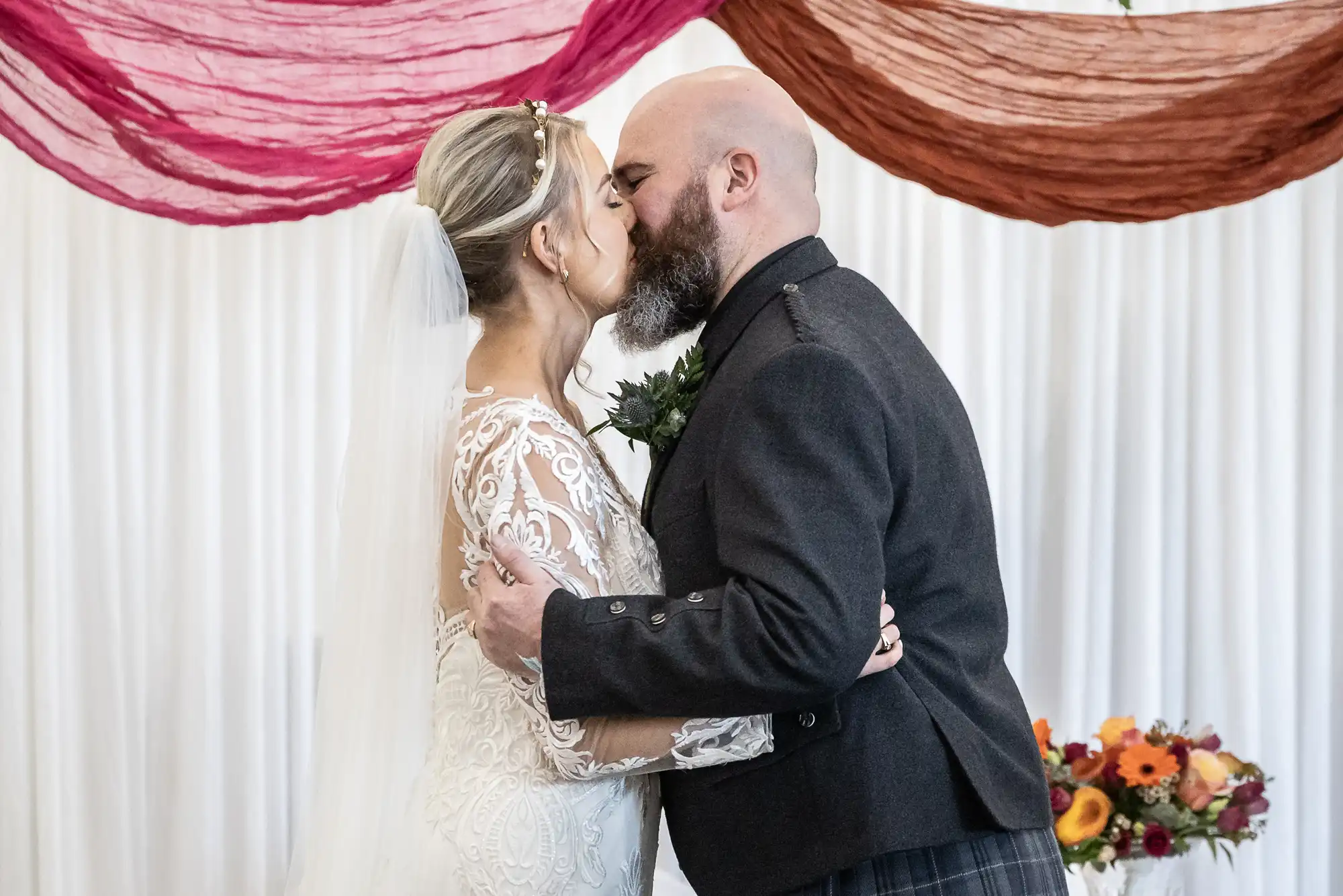 A bride and groom, both with closed eyes, kiss at their wedding. The bride wears a white lace dress and veil; the groom wears a dark suit with a buttonhole. Colourful drapes and a bouquet of flowers decorate the background.