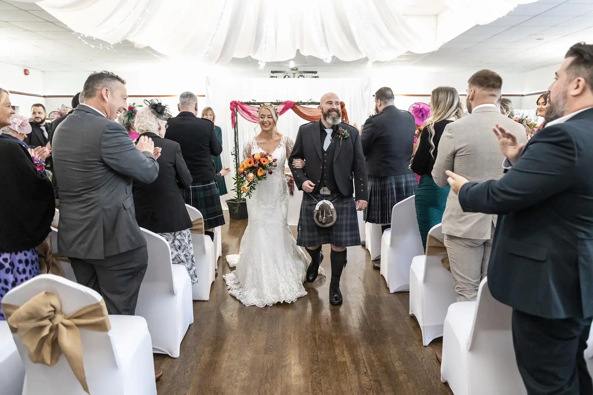 A smiling bride in a lace gown holding orange flowers walks arm-in-arm with a man in a kilt down an aisle lined with applauding guests at an indoor wedding ceremony, under white drapes and colourful decorations.