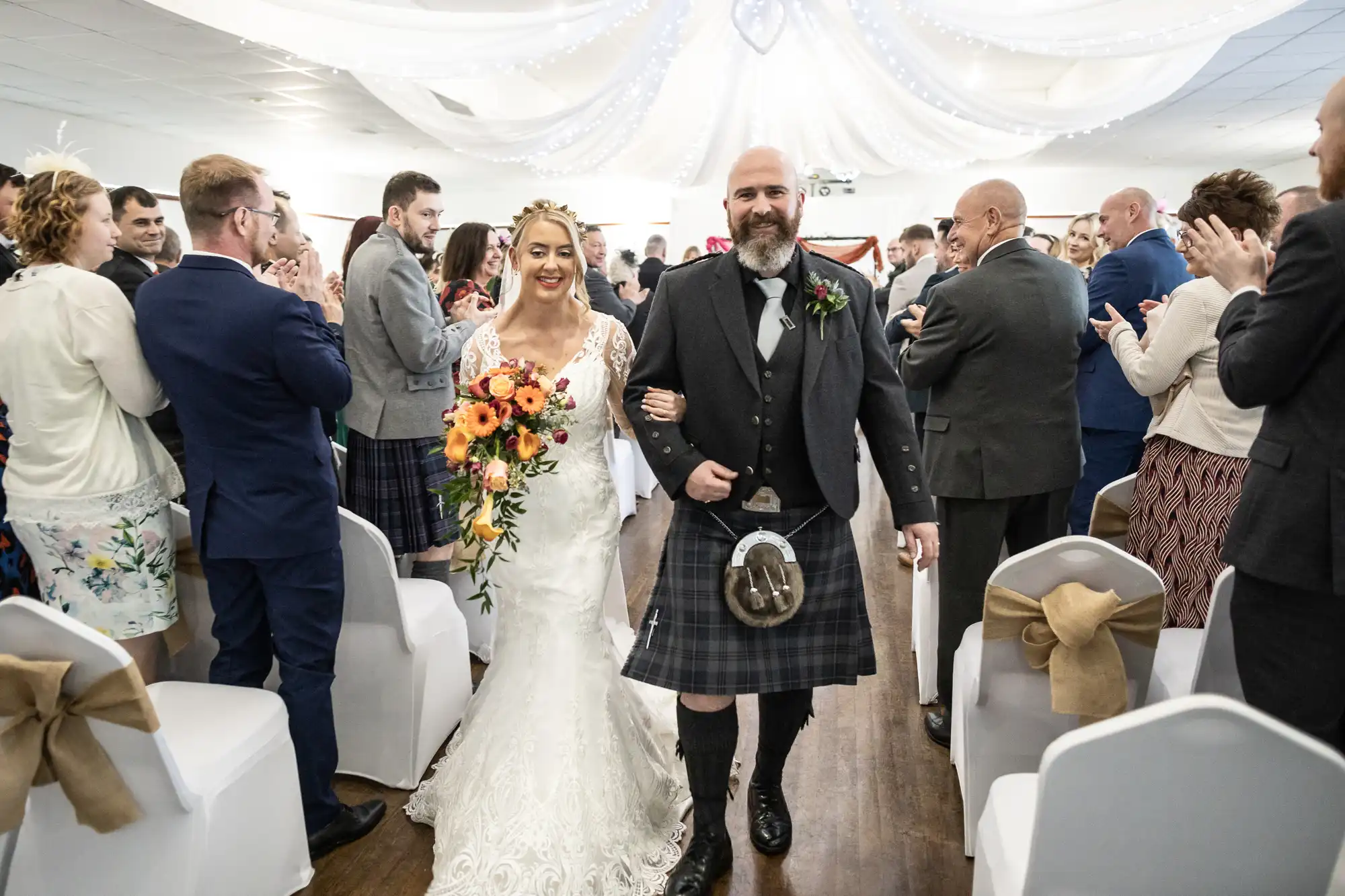 A smiling bride in a lace white gown and a groom in a black jacket and kilt walk down the aisle together, arm in arm, as wedding guests dressed formally stand and clap on either side in a decorated indoor venue with white drapes and lights above.