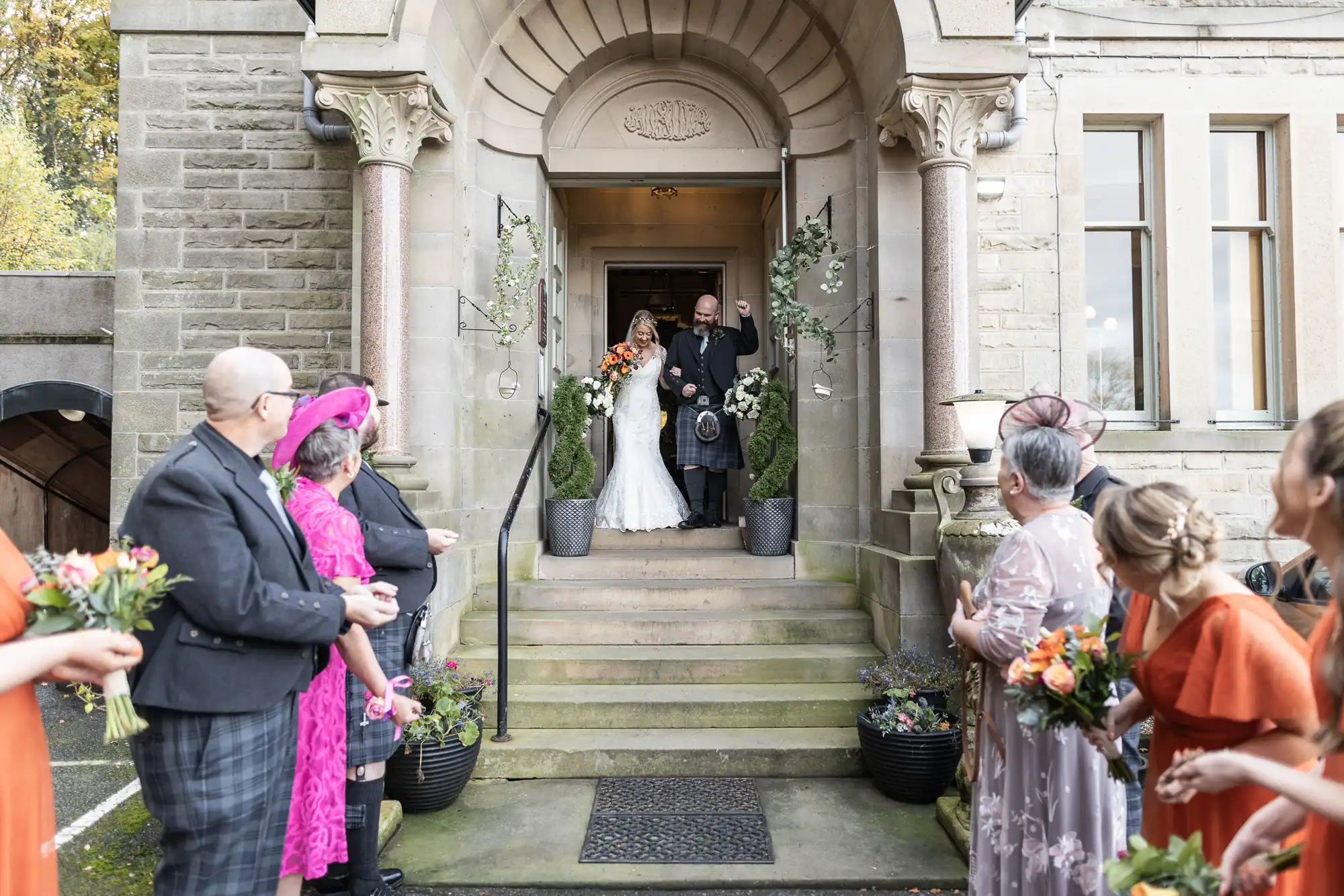 A bride in a white dress and a man in a kilt stand at the top of stone steps outside a grand building, smiling. Wedding guests holding bouquets stand on either side of the steps, wearing colourful outfits and looking towards the couple.