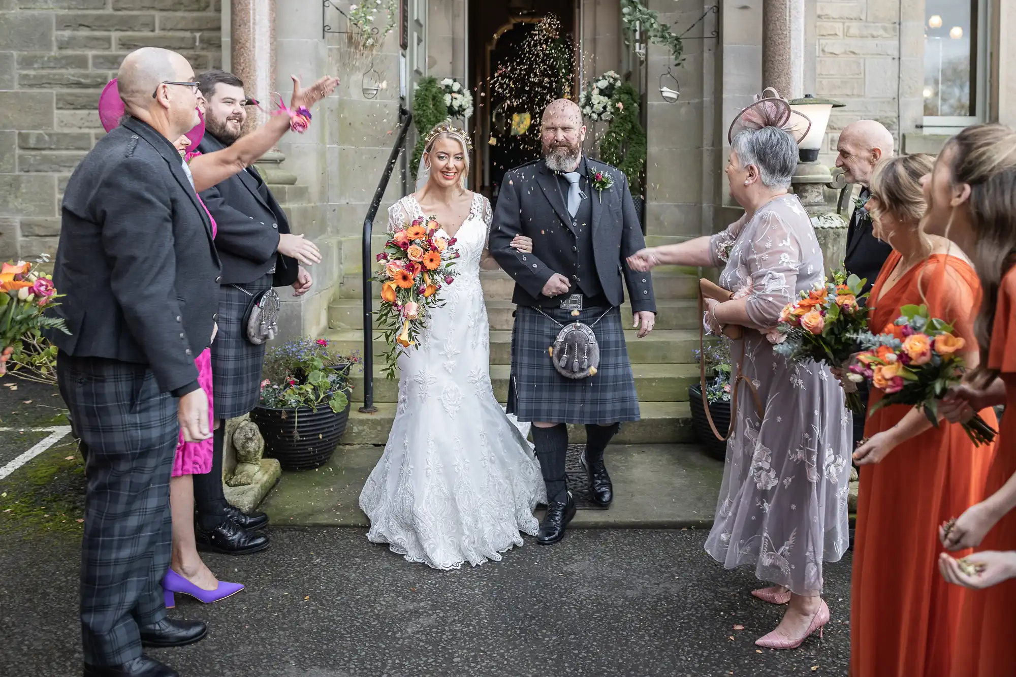 A bride in a white dress walks arm in arm with a man in a kilt outside a stone building, smiling. Guests in colourful outfits throw confetti and hold bouquets. Two large flower pots flank the doorway, and petals float in the air amid celebration.