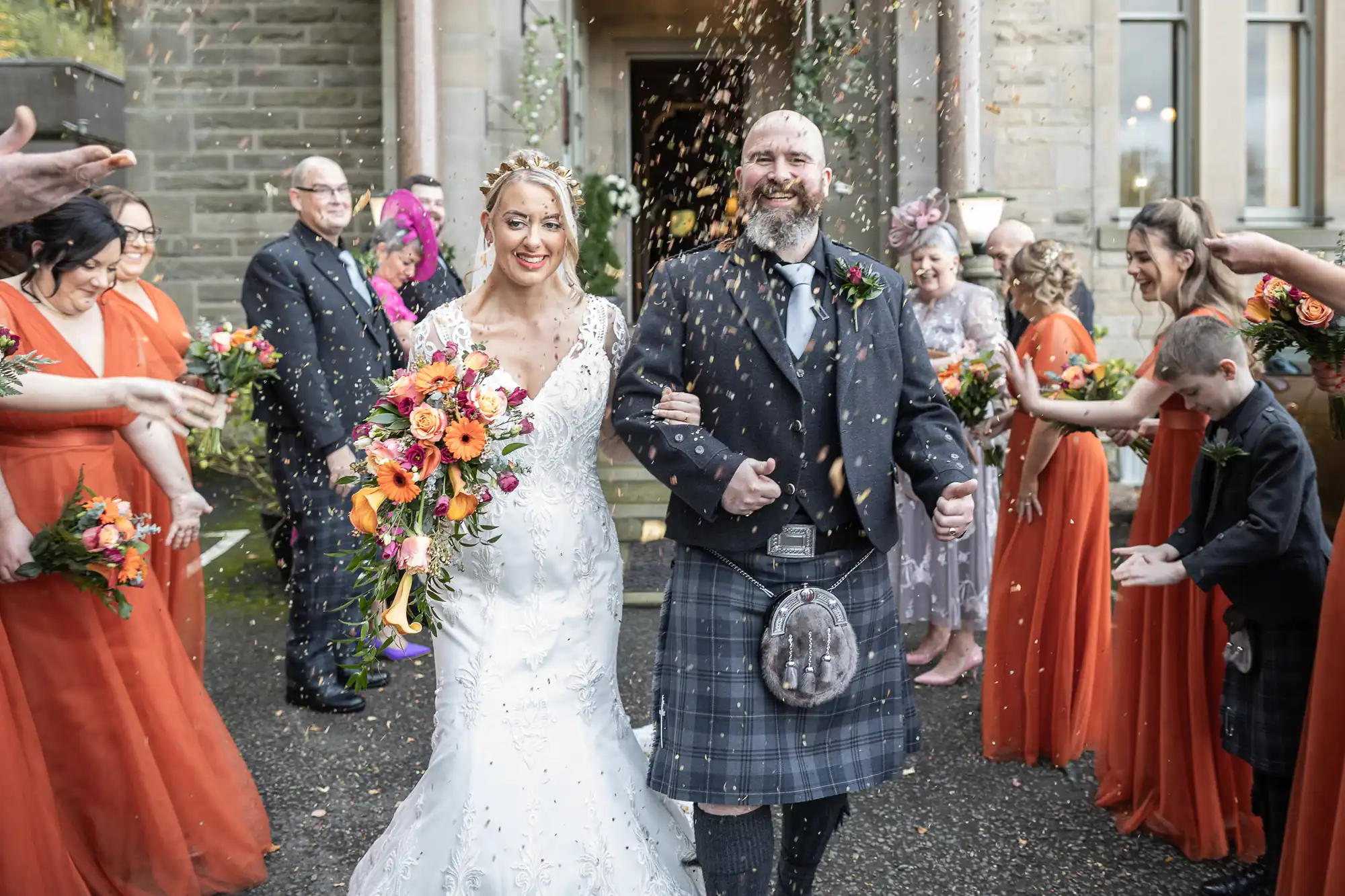 A smiling bride in a white dress and a groom in a kilt walk arm in arm as guests in orange dresses throw confetti. The couple hold bouquets and are surrounded by people celebrating outside a stone building.