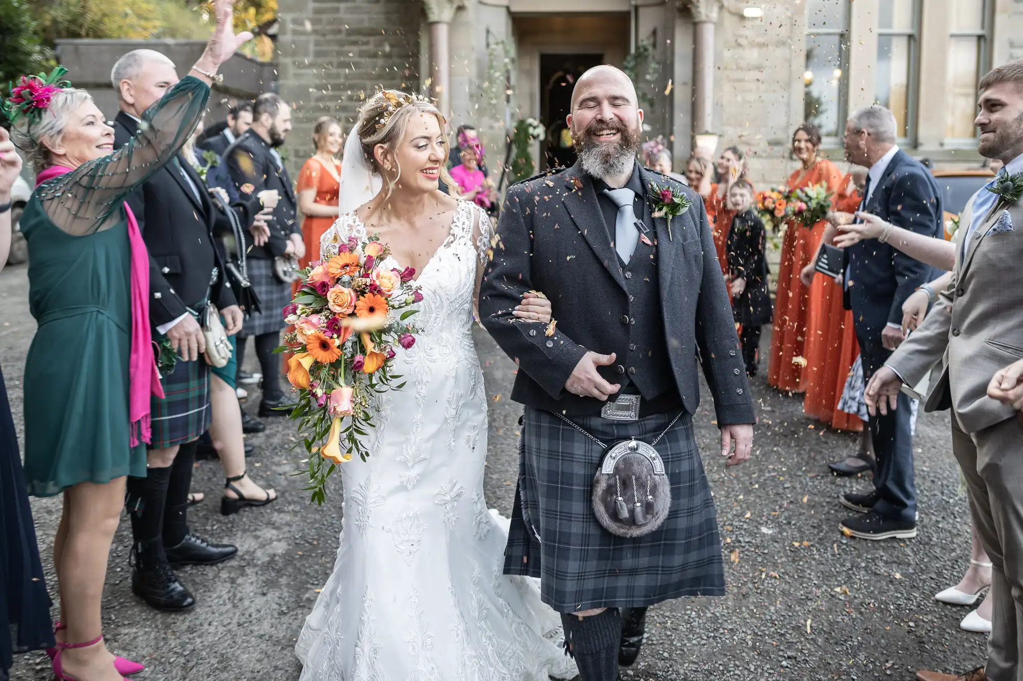 A smiling bride in a white lace gown and a groom in a grey kilt walk arm-in-arm outside, surrounded by guests. The bride holds a vibrant bouquet; guests throw confetti and cheer. Everyone looks joyful, dressed in formal attire, with greenery in the background.