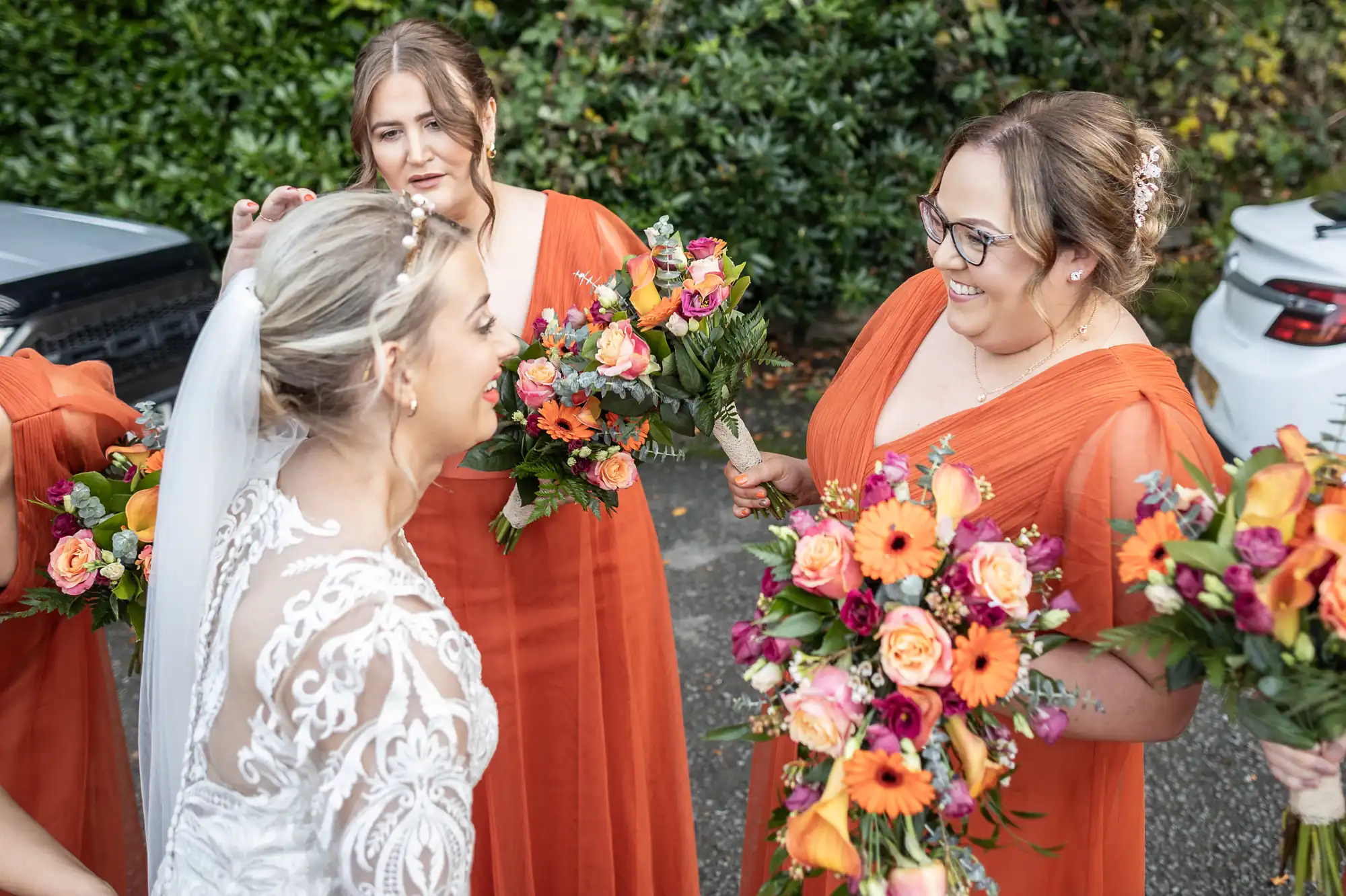 A bride in a lace wedding dress smiles at two bridesmaids in orange dresses holding vibrant bouquets of orange and pink flowers, standing outdoors near green foliage and parked cars.