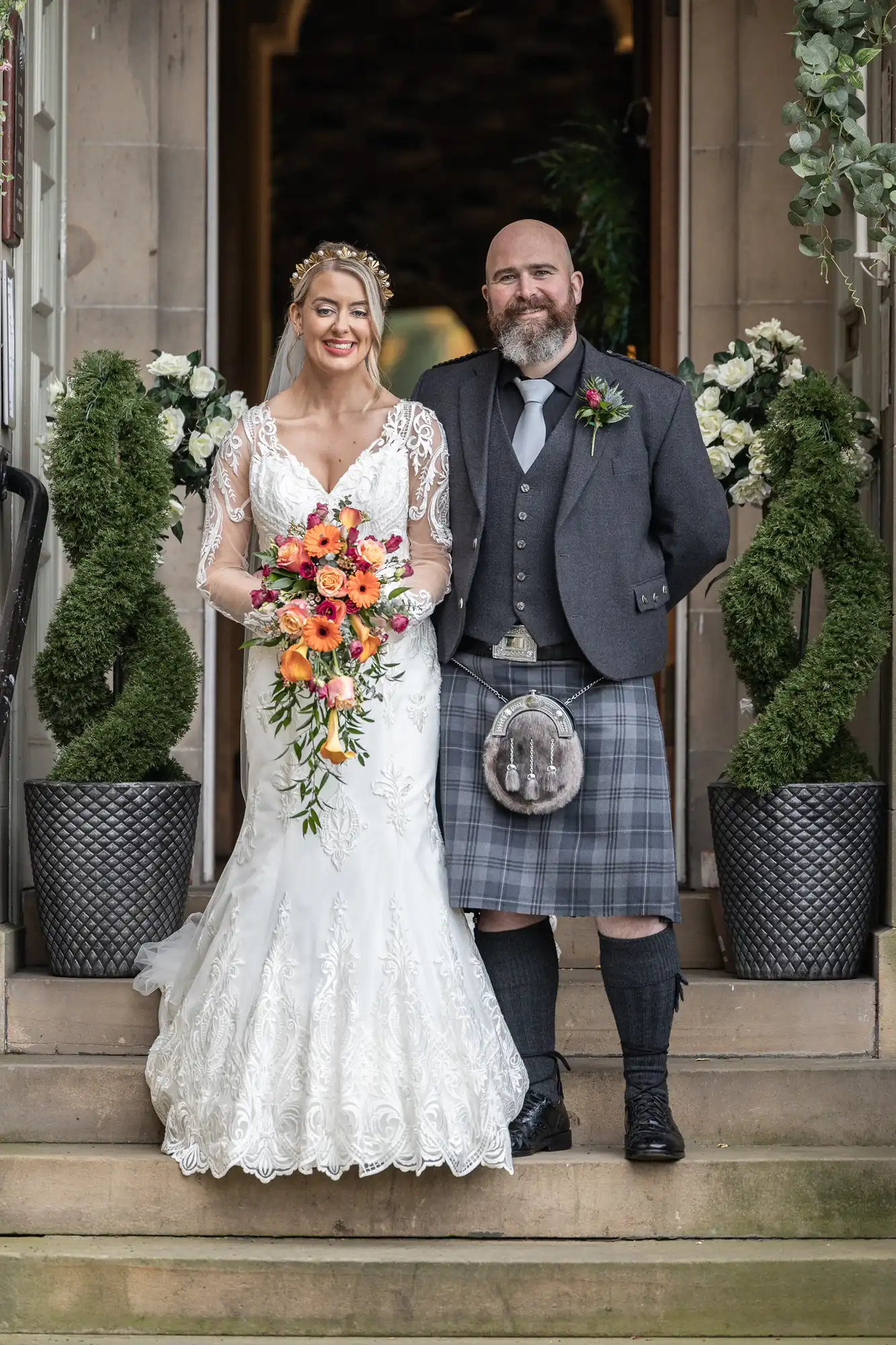 A bride in a white lace gown holds a vibrant bouquet and stands beside a groom in a grey jacket, plaid kilt, and sporran. Both smile, posed on stone steps, with spiral-shaped topiary and flower arrangements in the background.
