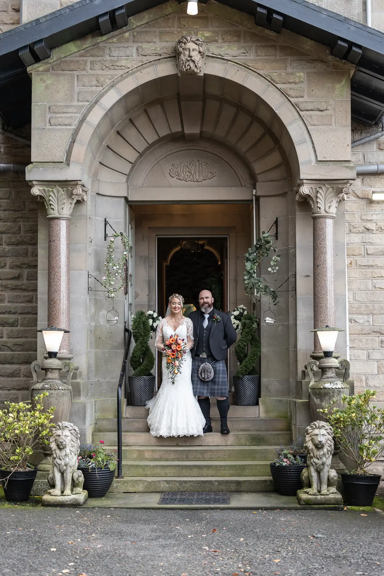A bride in a white dress holding a bouquet stands beside a groom in a kilt on stone steps under an ornate arched entrance. The doorway is decorated with greenery in pots, and stone lion statues guard the steps. The building has stone walls and columns.