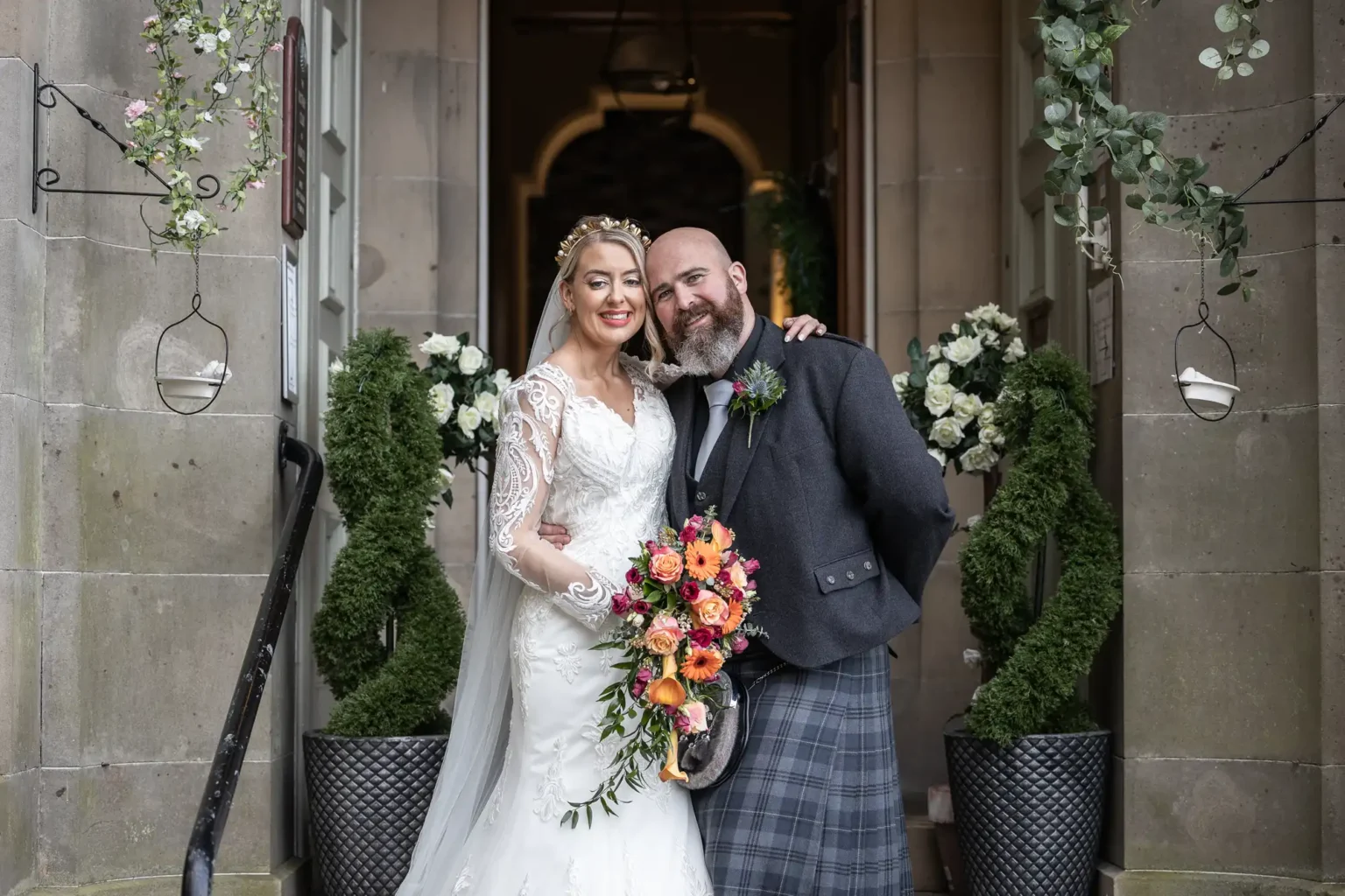 Mansfield House Hotel wedding photographer image of a bride in a white lace dress and veil smiles, holding a colourful bouquet, standing beside a bearded groom in a dark jacket and tartan kilt. They pose at an archway decorated with greenery and white flowers, flanked by potted spiral topiary plants.