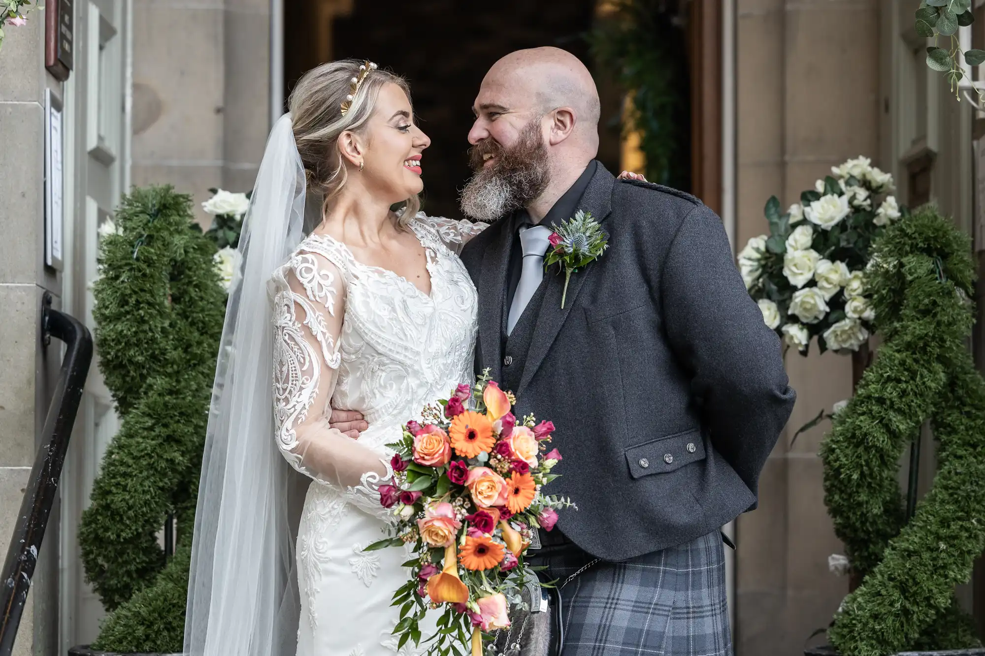 A smiling bride in a white lace dress and veil stands arm-in-arm with a bearded groom in a grey jacket and checked kilt, holding a vibrant bouquet of orange, pink, and white flowers, surrounded by greenery and white floral arrangements at a wedding venue entrance.