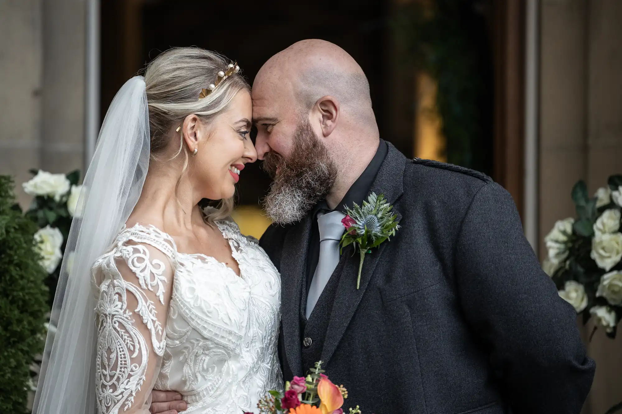 A bride in a white lace dress and veil smiles, touching foreheads with a bearded groom in a dark suit and buttonhole. They stand close, holding a vibrant bouquet, surrounded by white flowers and greenery, looking happy and in love.