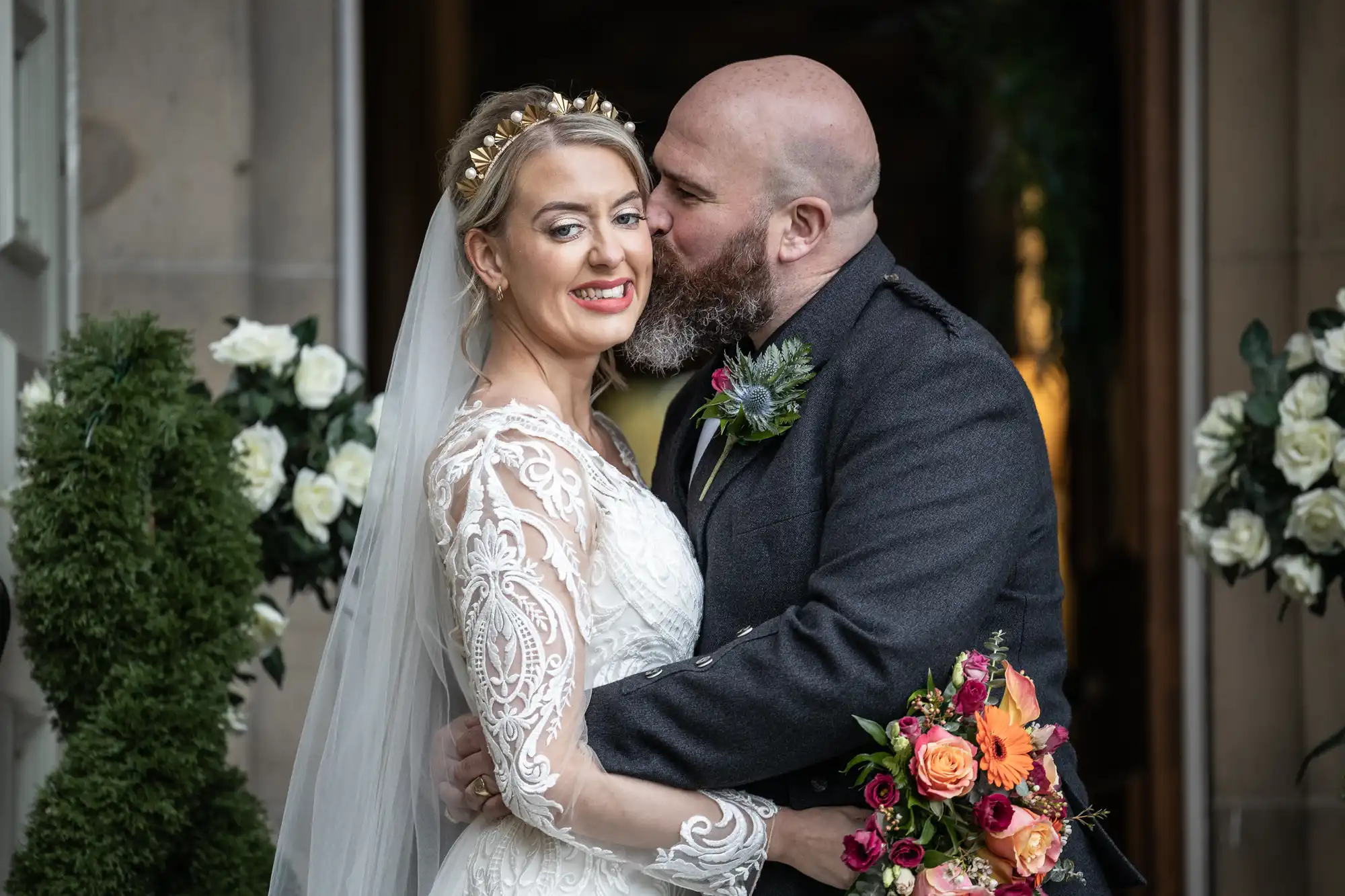 A bride in a white lace dress and veil smiles whilst being kissed on the cheek by a bearded groom in a dark suit. She holds a colourful bouquet of flowers. White roses and greenery decorate the background, creating a romantic setting.