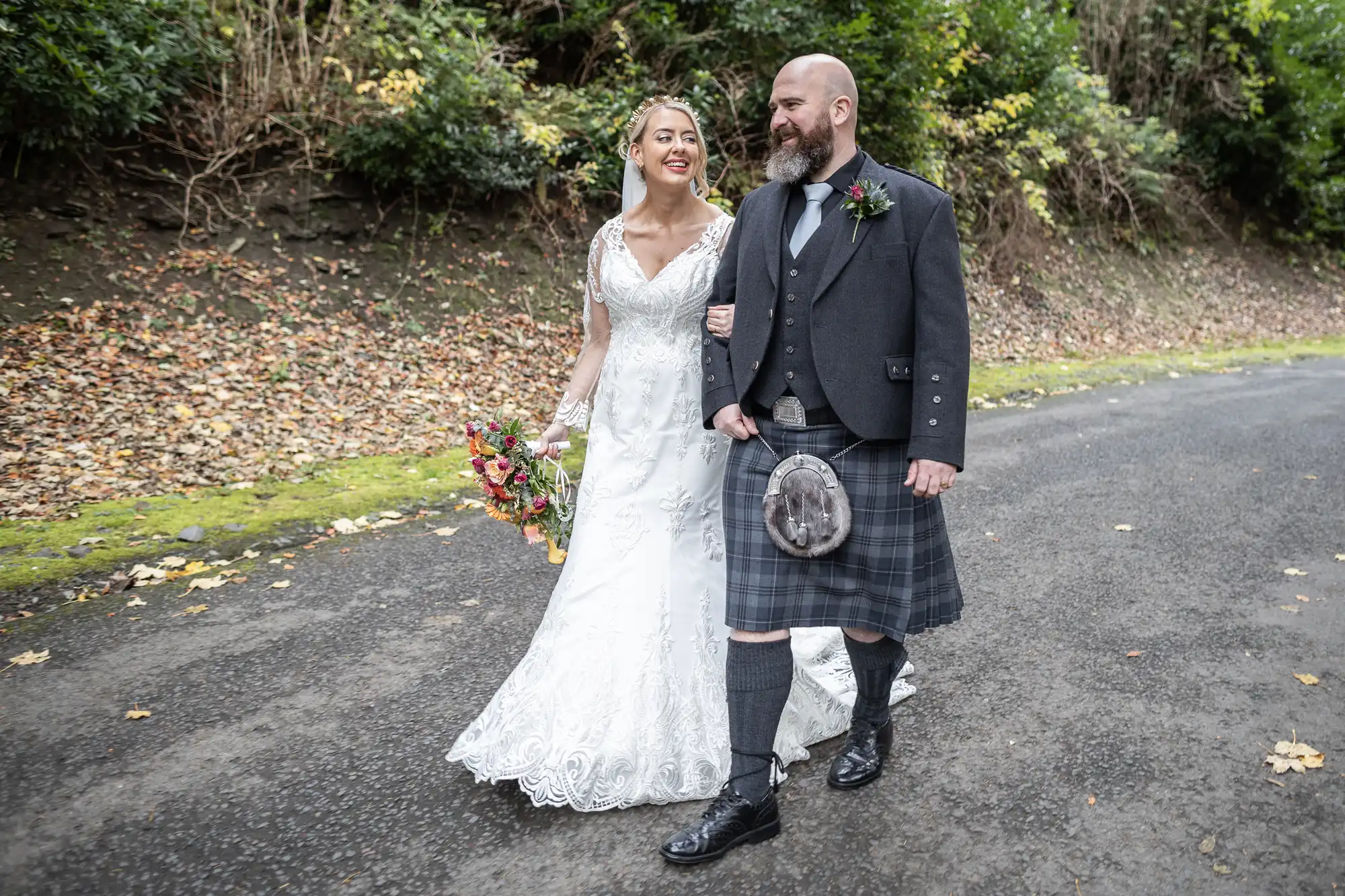 A bride in a white lace gown holds a bouquet and smiles at a groom in a kilt and jacket as they walk arm-in-arm down a paved path, surrounded by autumn leaves and greenery, both appearing happy and relaxed outdoors.