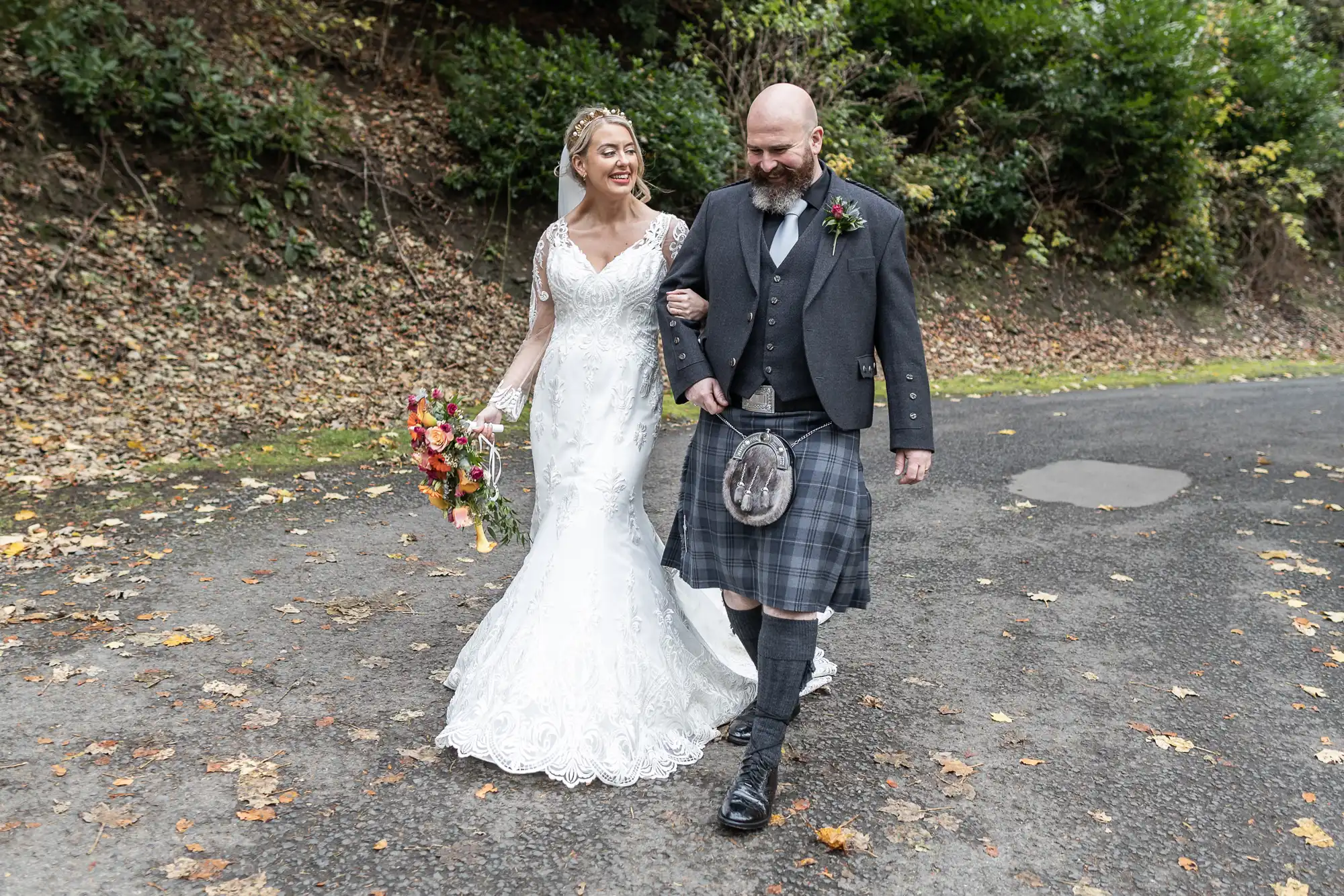 A smiling bride in a white lace gown and veil walks arm-in-arm with a bearded groom in a dark jacket, kilt, and sporran. The bride holds a bouquet of flowers. They walk on a leaf-strewn path with greenery in the background.