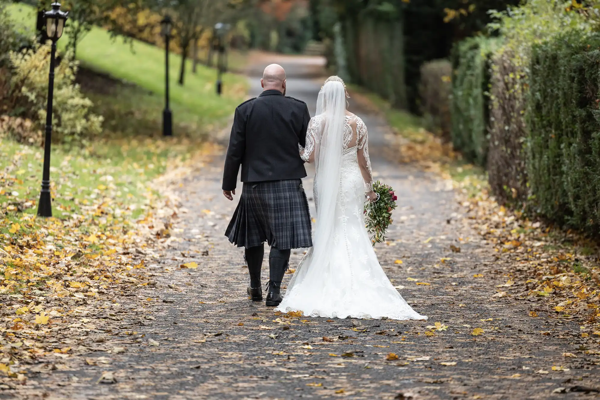 A bride in a white dress and veil walks arm-in-arm with a groom in a black jacket and kilt down a leaf-covered path outdoors, surrounded by greenery and autumn trees. The couple is seen from behind, holding a bunch of flowers.