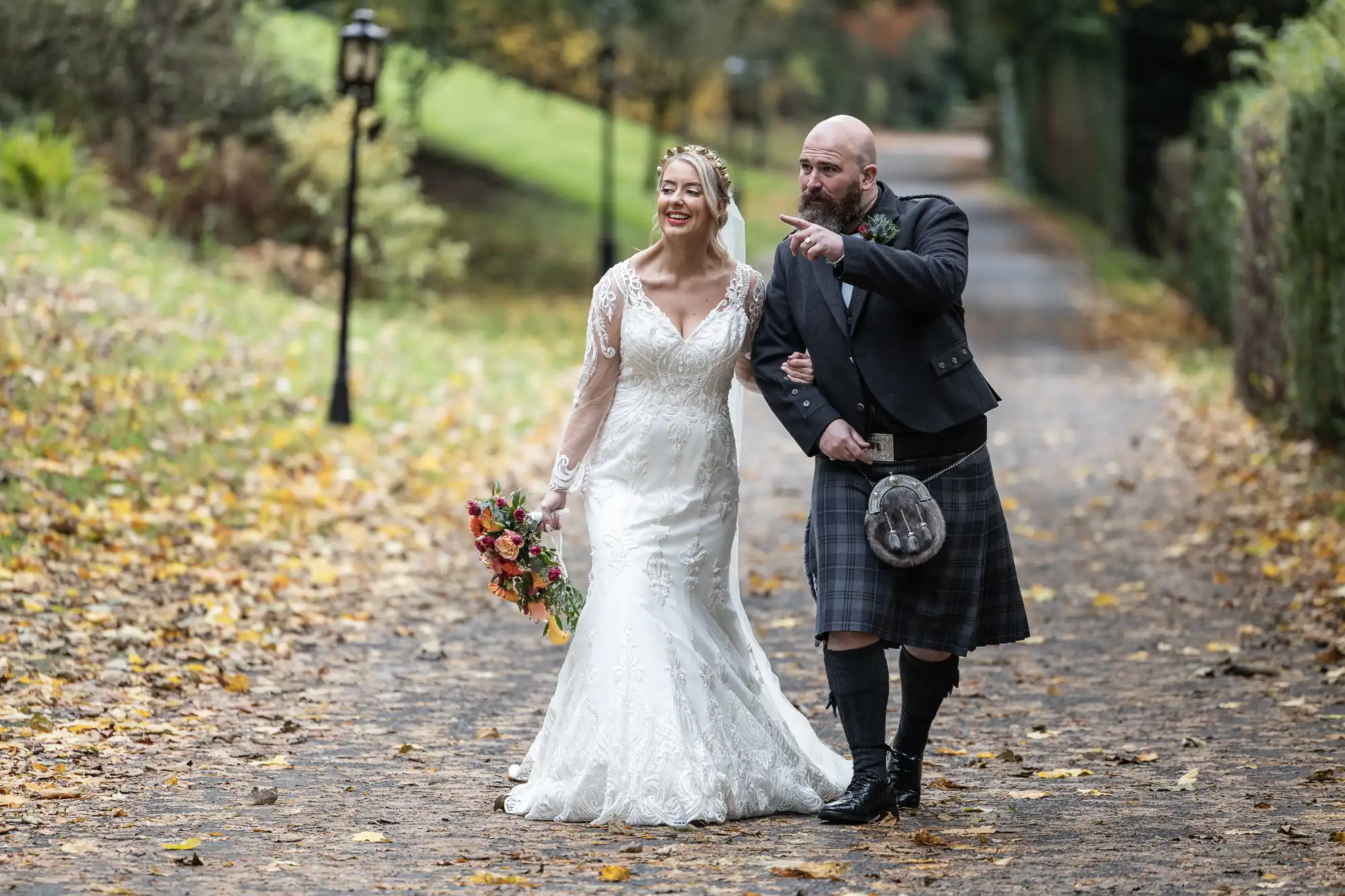 A bride in a white lace gown holding a bouquet walks arm in arm with a man in a traditional Scottish kilt and jacket. They walk on a leaf-strewn path, smiling; the man gestures ahead whilst autumn trees and a lamp-post frame the scenic background.