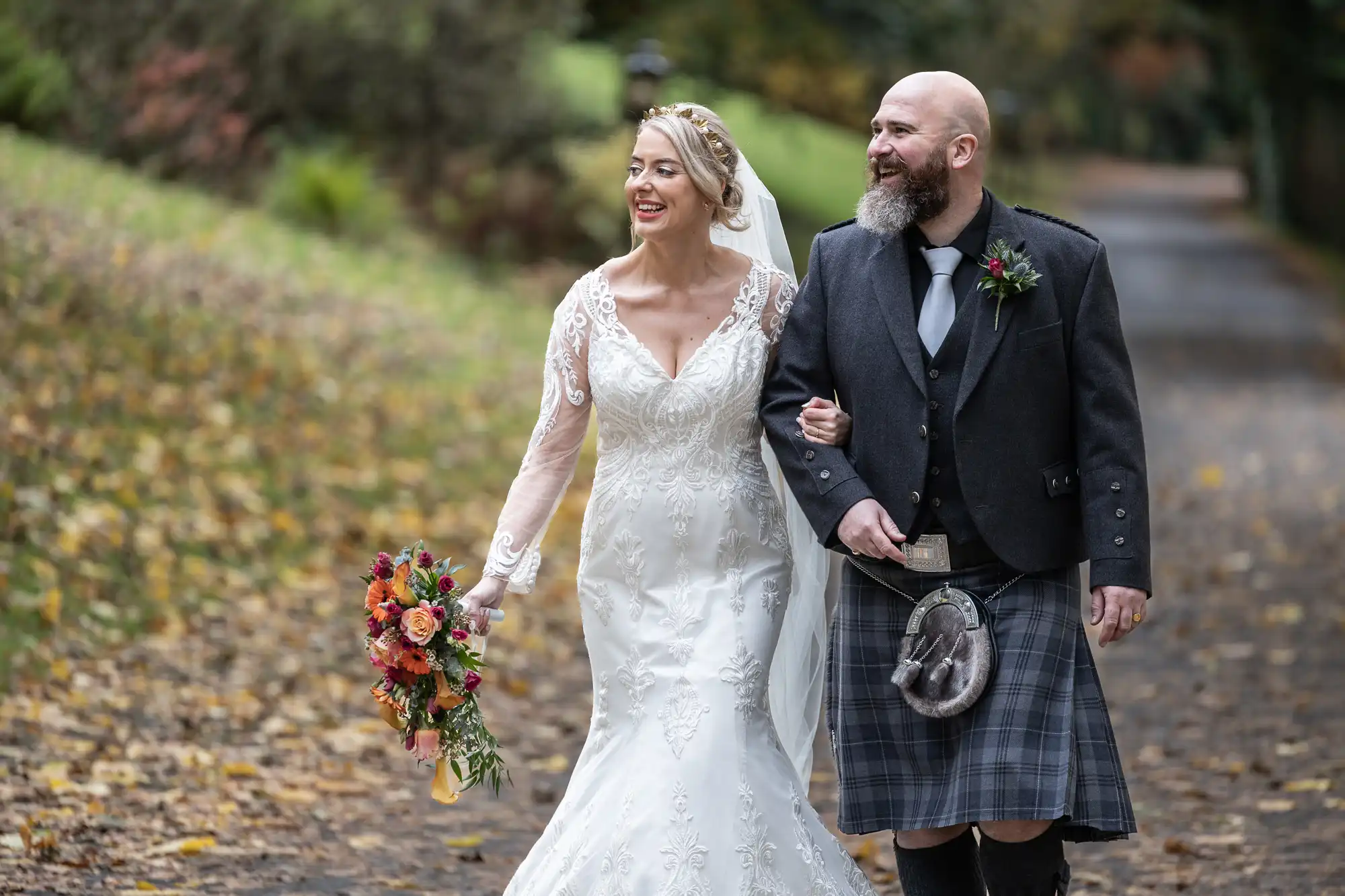 A smiling bride in a white lace gown and veil walks arm-in-arm with a bearded groom in a dark jacket and traditional tartan kilt. She holds a colourful bouquet. They walk outdoors on a leaf-strewn path with greenery in the background.