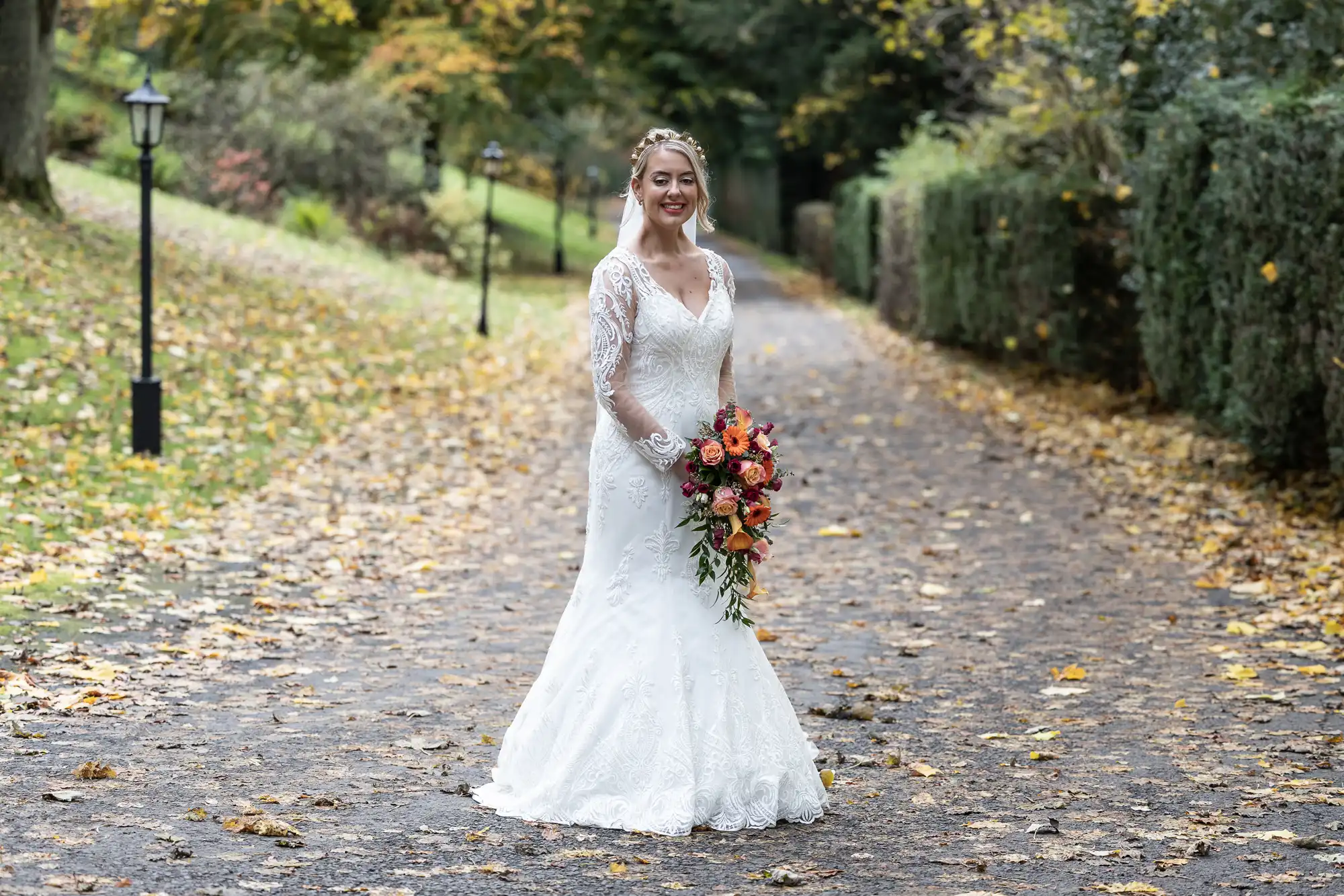 A bride in a white lace gown holds a vibrant bouquet of orange and red flowers. She stands smiling on a leaf-covered path lined with green hedges and autumn trees, with soft natural light highlighting the peaceful outdoor setting.