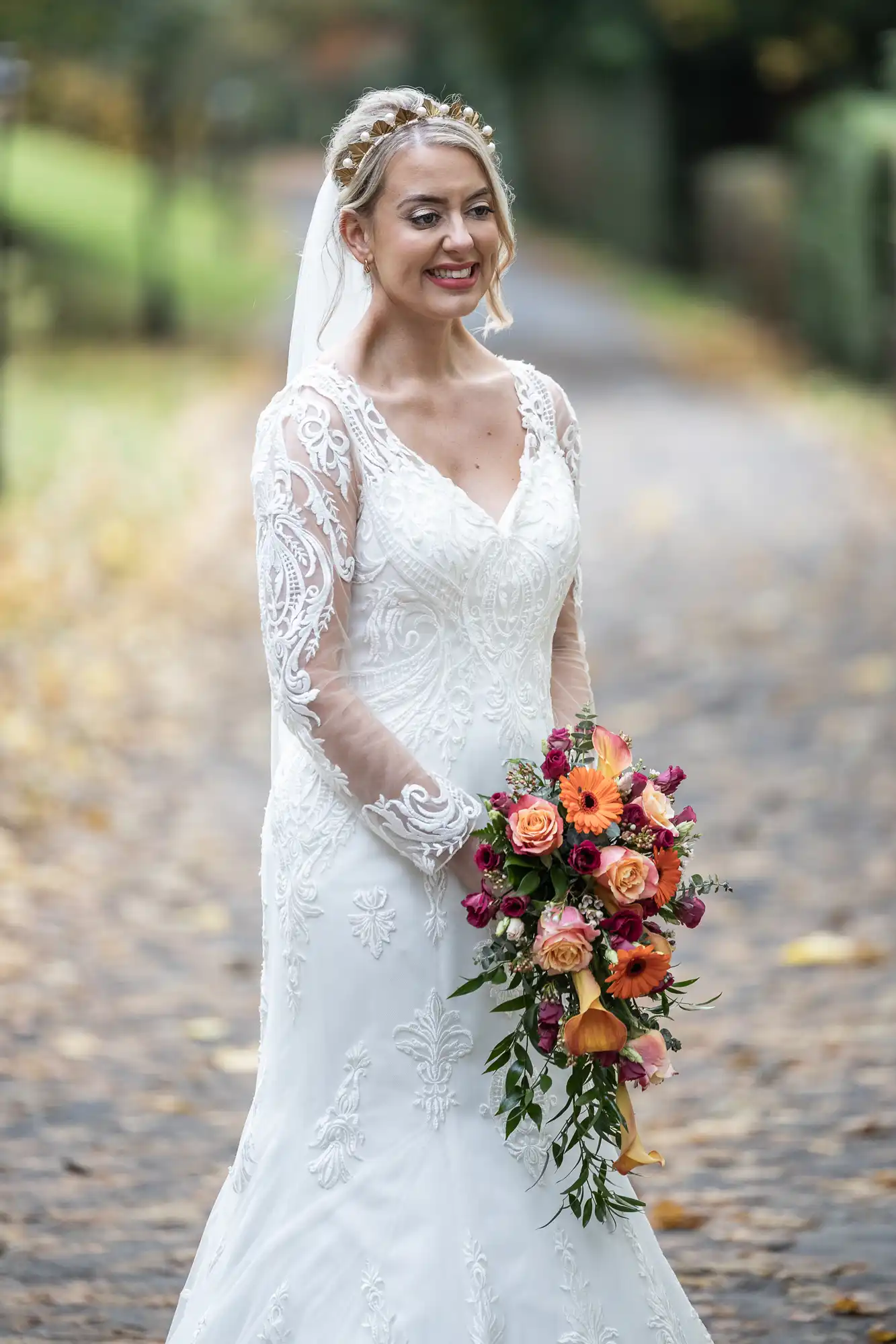 A smiling bride stands outdoors on a leafy path, wearing a white, long-sleeved lace wedding dress and veil. She holds a cascading bouquet of orange, pink, and red flowers with greenery. Trees and a blurred background suggest an autumn setting.