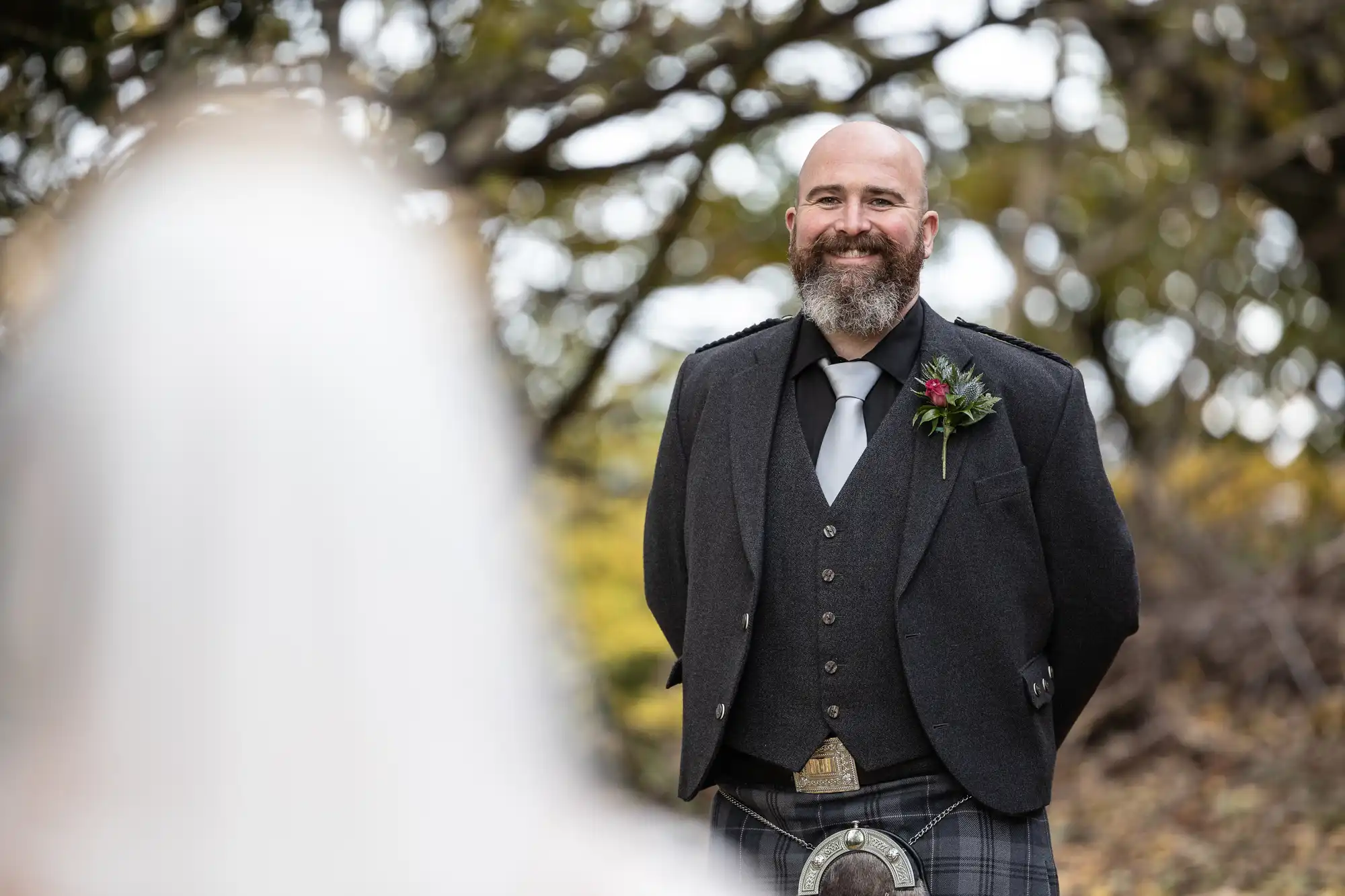 A bearded man in formal Scottish attire, including a dark jacket, waistcoat, tie, and kilt, smiles warmly outdoors. A blurred figure in white, likely a bride, is in the foreground. Trees with green leaves fill the background.