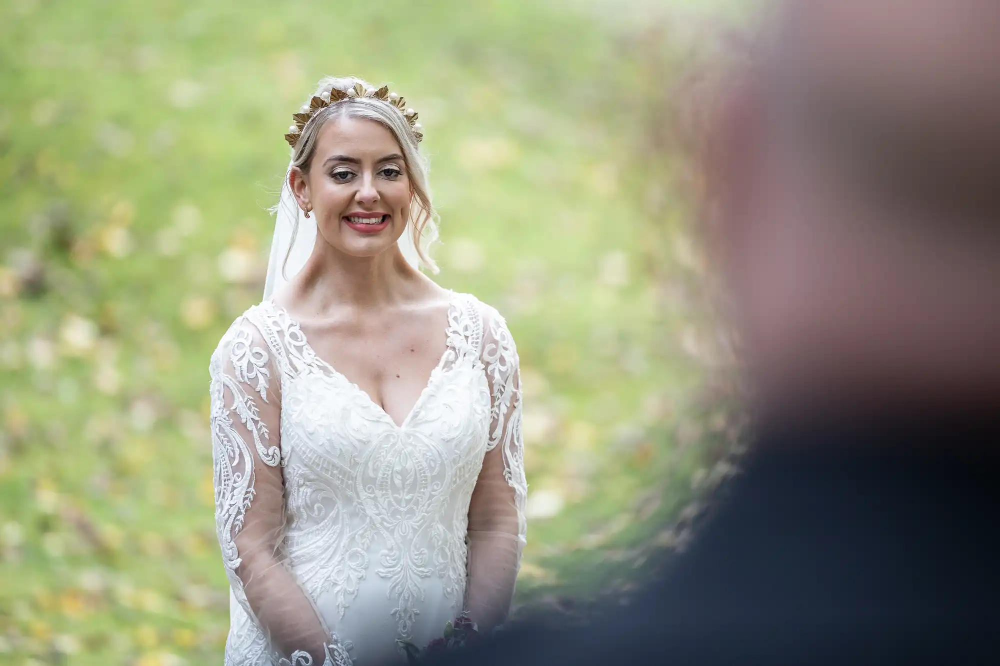 A bride in a white lace wedding dress and gold floral headpiece smiles outdoors. She stands on green grass with scattered yellow leaves, looking towards a blurred figure in the foreground. The background is softly focussed, emphasising the bride’s expression.