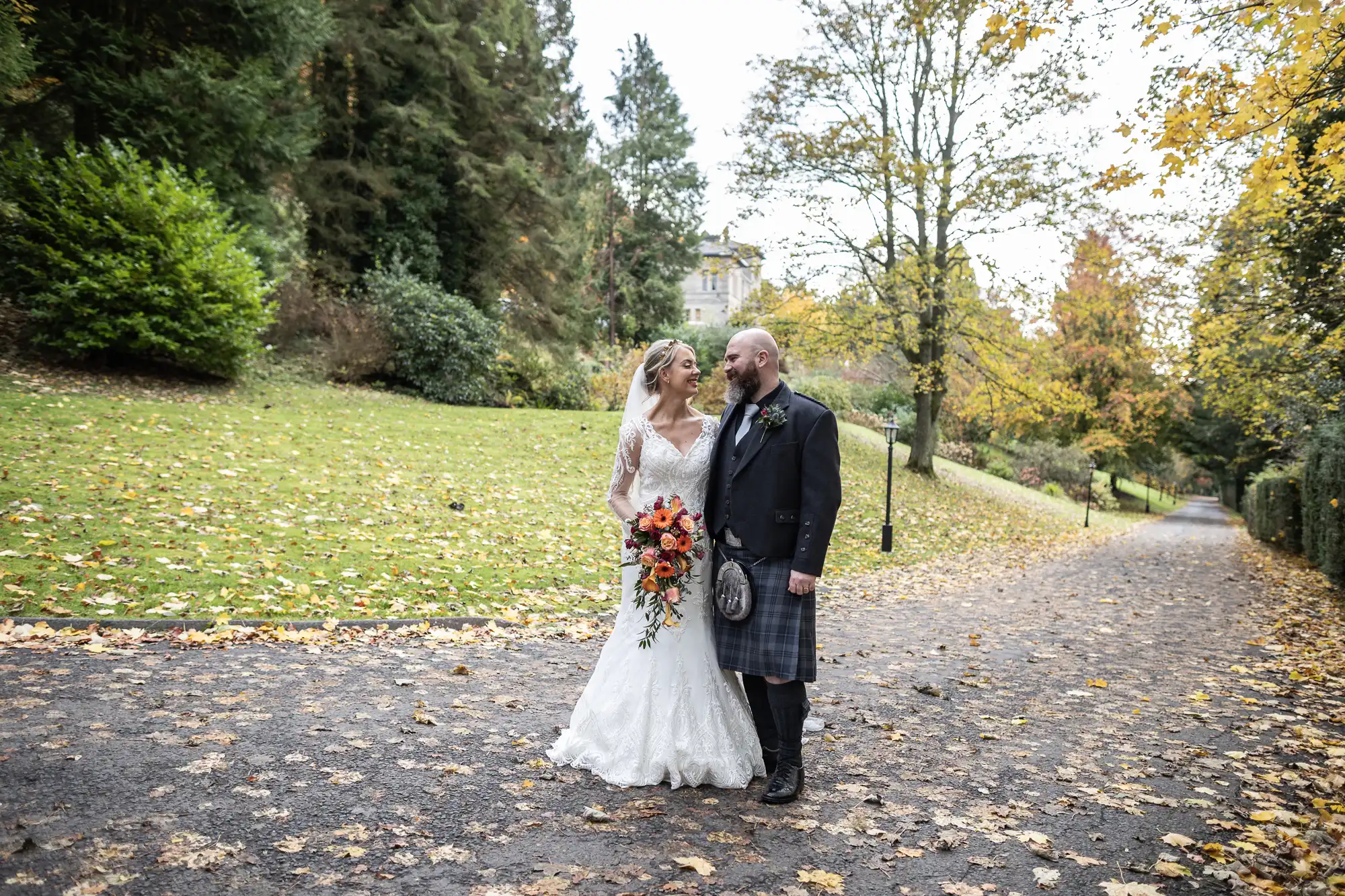 A bride in a white lace gown and a groom in a Scottish kilt stand close together on a leaf-strewn path, surrounded by green lawn and autumn trees, gazing at each other with a bouquet of orange flowers in the bride’s hand.