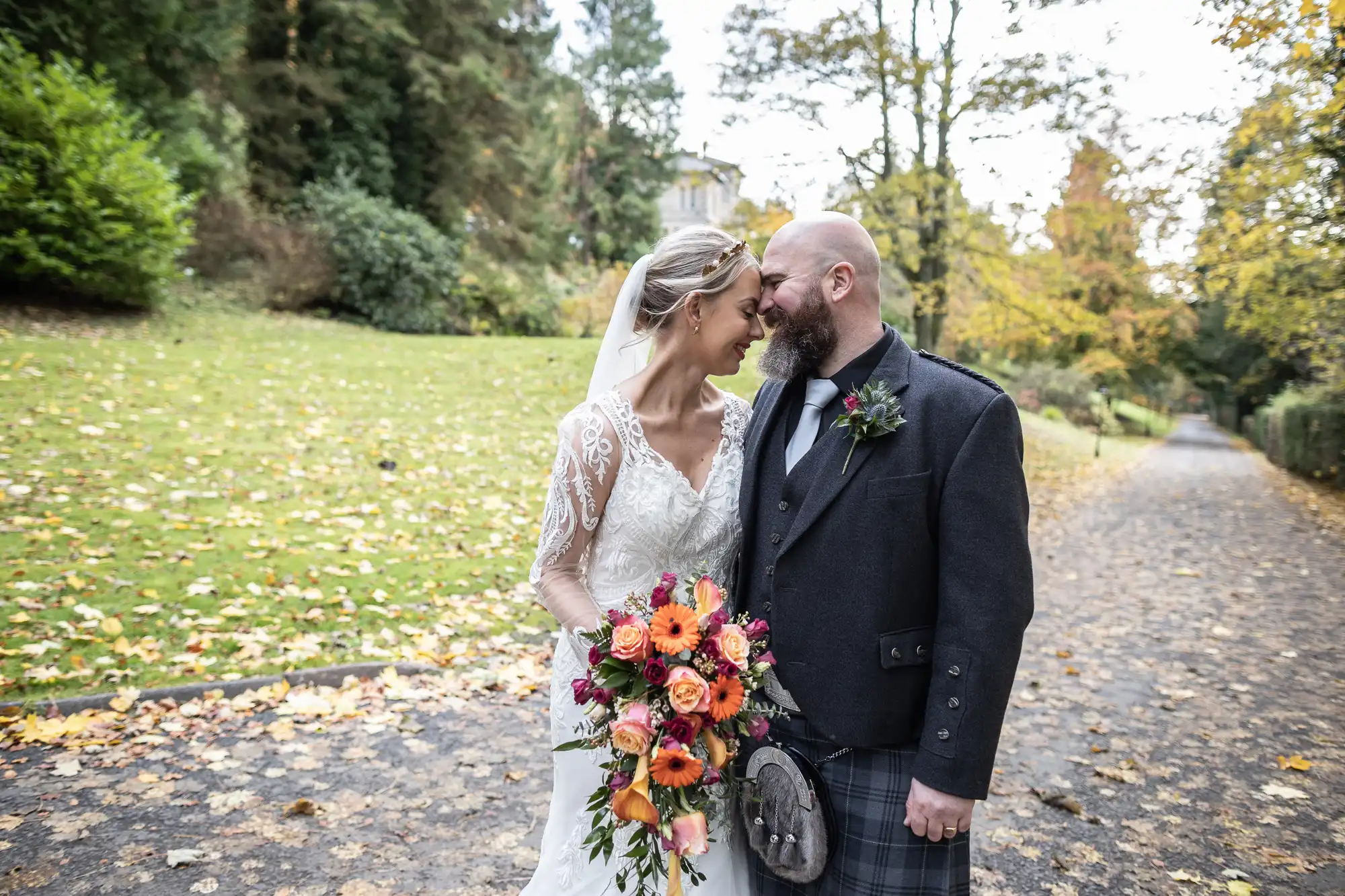 A bride in a white lace gown and veil holds a colourful bouquet as she smiles, touching foreheads with a bearded groom in a dark jacket and tartan kilt. They stand on a tree-lined path covered in autumn leaves, with greenery and soft sunlight in the background.