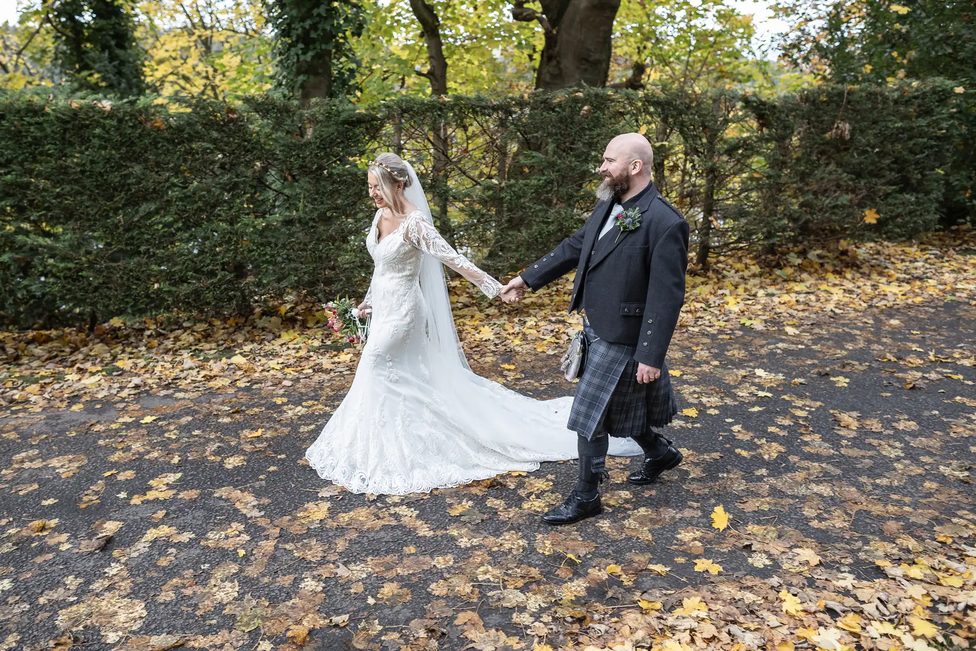 A bride in a long white lace gown and veil holds hands with a groom in a black jacket and traditional tartan kilt as they walk outside on a leaf-covered path, surrounded by autumn trees and a hedge.