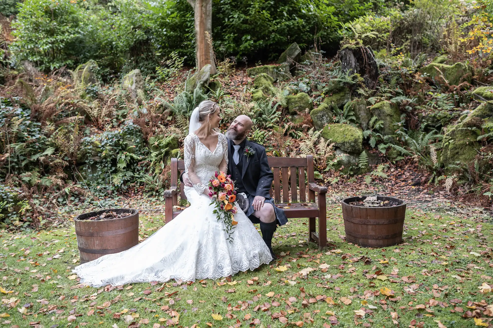 A bride in a white dress and veil sits beside a groom in a dark suit and kilt on a wooden bench in a lush, leafy garden. The bride holds a vibrant bouquet. Both smile at each other, surrounded by ferns, moss, and two wooden barrels.
