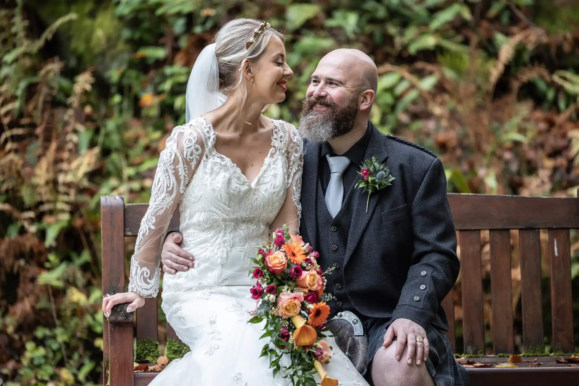 A bride in a white lace wedding dress and veil sits smiling beside a bearded groom in a dark kilt jacket on a wooden bench. The bride holds a vibrant bouquet of orange, red, and yellow flowers. Green foliage and autumn leaves fill the background.
