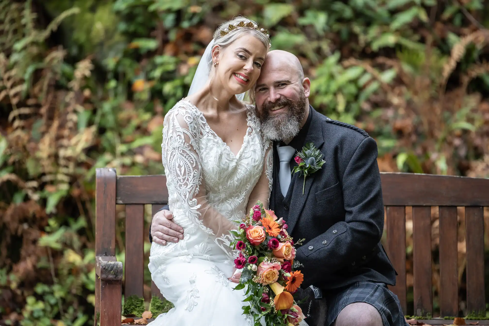 A smiling bride in a white lace dress and a groom in a dark jacket and kilt sit close together on a wooden bench, surrounded by greenery and autumn leaves. The bride holds a colourful bouquet of orange and pink flowers.