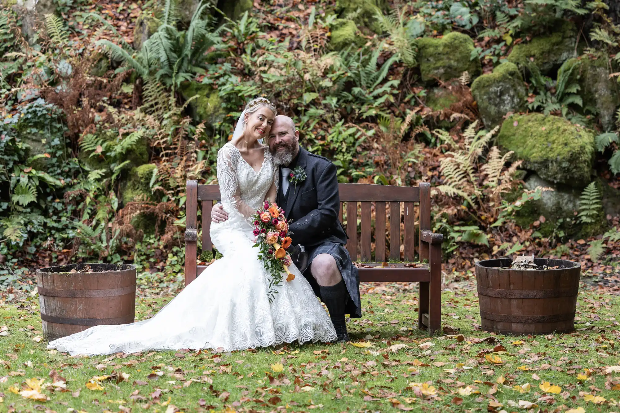 A bride in a white lace dress and a groom in a kilt sit smiling on a wooden bench outdoors. The bride holds a colourful bouquet. Ferns and moss-covered rocks fill the background, and two wooden barrels sit on either side of the couple.