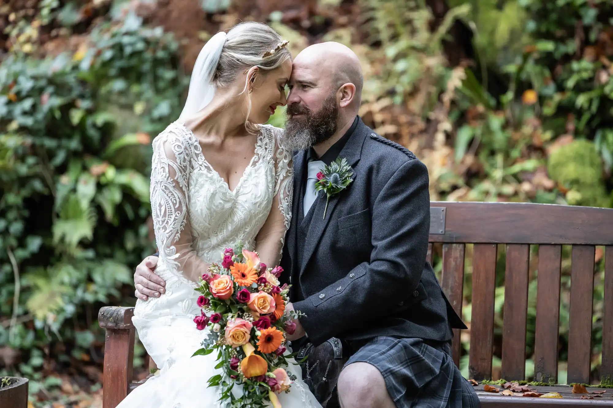 A bride in a lace wedding dress and veil sits on a bench, holding a colourful bouquet of roses and greenery. She smiles and leans her forehead against a bearded groom in a dark kilt and jacket, surrounded by lush greenery outdoors.