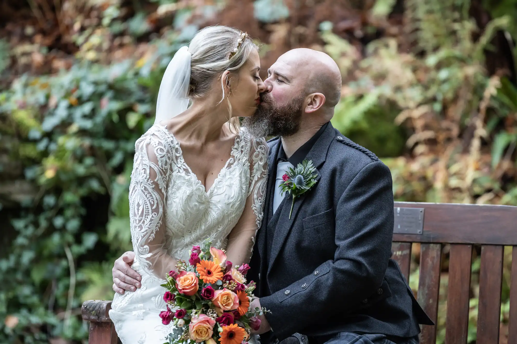 A bride in a white lace dress and veil sits closely with a bearded groom in a dark suit as they share an intimate kiss on a wooden bench. The bride holds a bouquet of colourful flowers. Lush greenery and foliage fill the background.